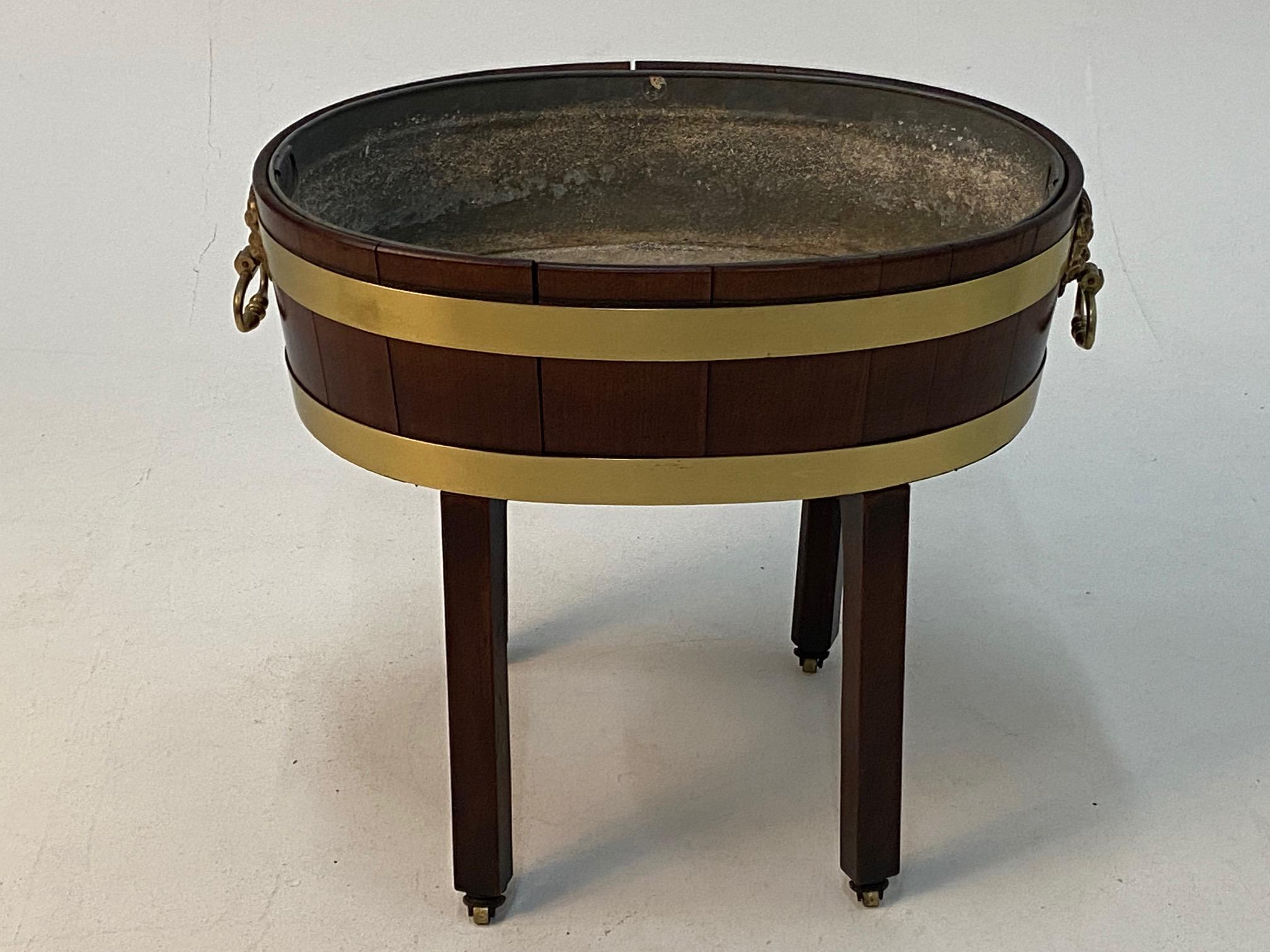 A beautiful antique English Georgian style oval mahogany cellarette on stand having handsome brass banding and figural handles with detailed lion or gargoyle-esque hardware. Legs terminate in brass casters and original metal liner is
