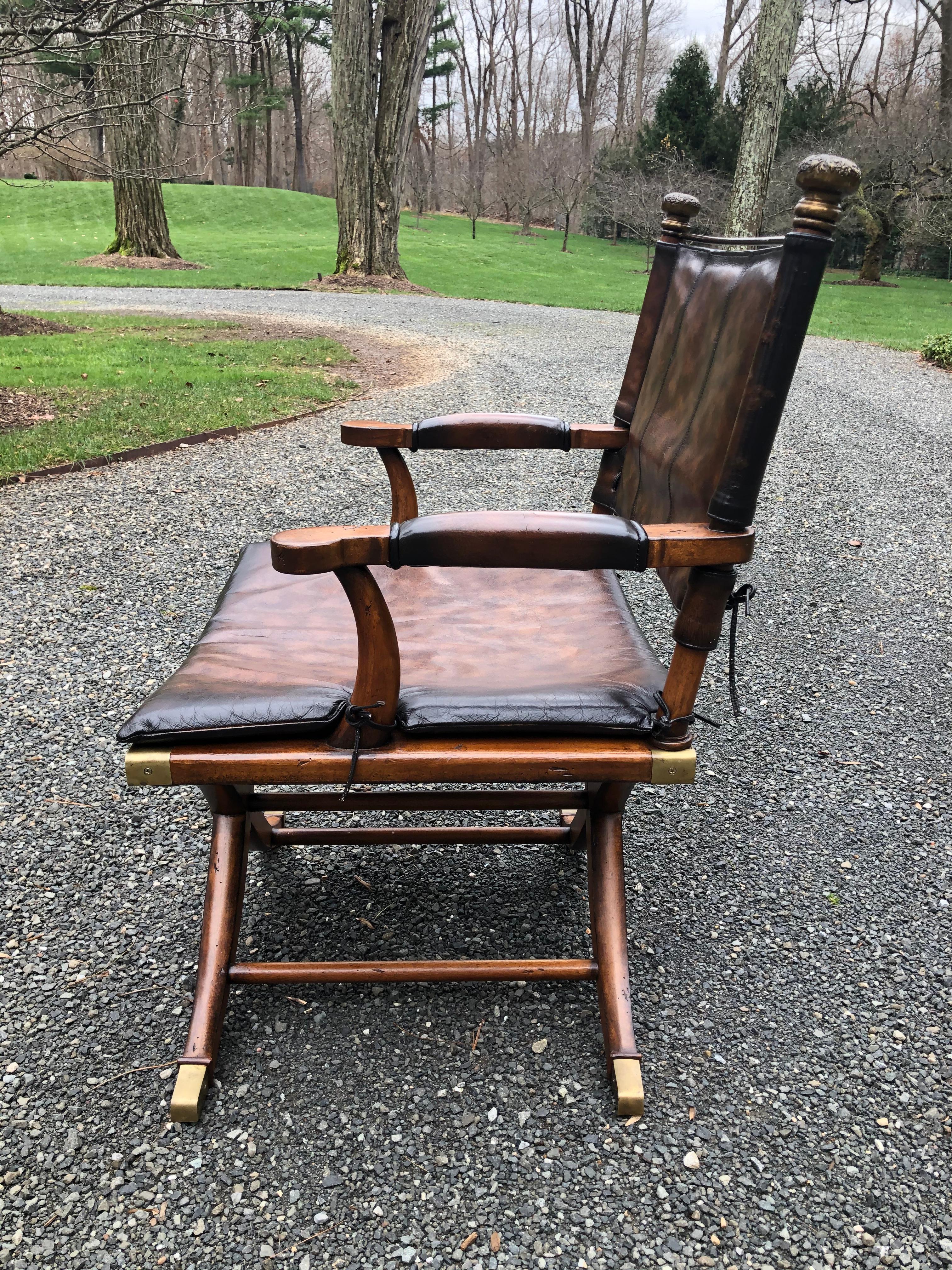 Handsome Campaign or safari chair in mahogany and leather. This chair has a leather back and seat cushion with ties; the seat underneath is wood and is beautifully decorated with antiqued brass mounts on the feet and the seat and has a medallion