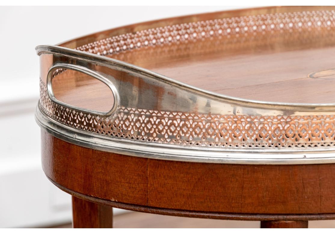 A finely crafted oval mahogany tray top table made in one piece. The top with a center fan inlay with ebonized details and a pierced copper tone silverplate gallery with inset twin handles. Raised on four curved tapering flared legs with an X form