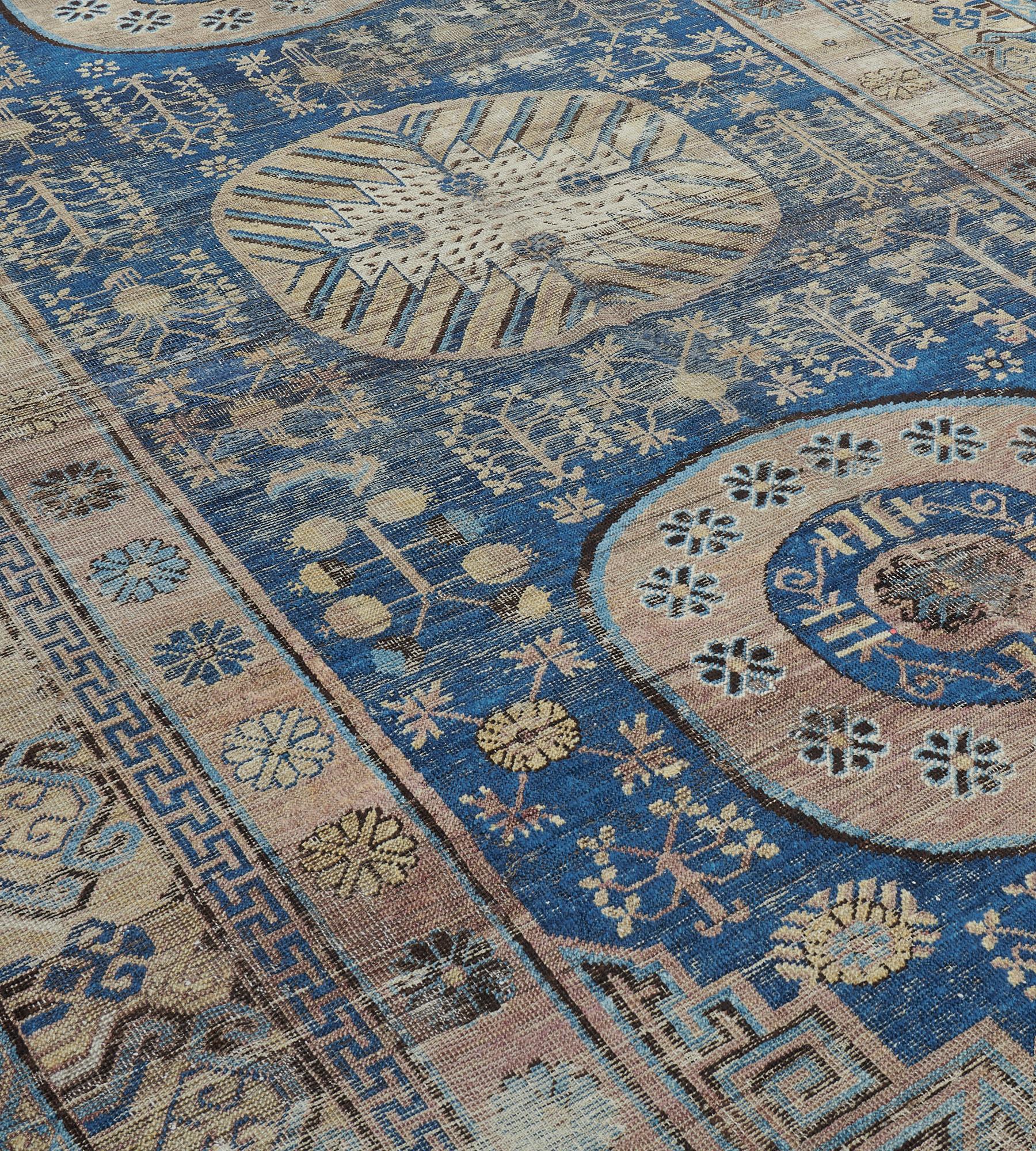 Ce tapis Khotan ancien présente un champ bleu roi ombragé parsemé de tiges fleuries, de têtes de fleurs et d'arbres feuillus autour d'une colonne de trois grands rondeaux, le central avec un grand losange cuspidé ivoire émettant des vrilles