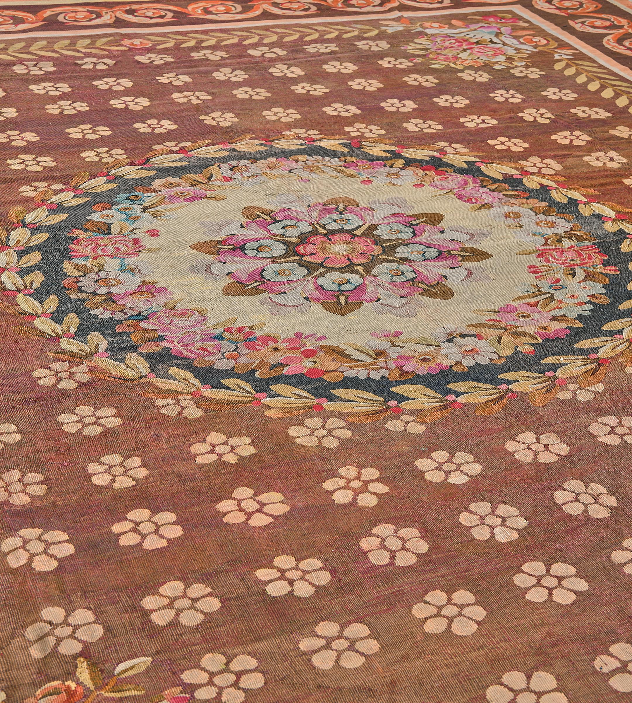 Questo antico tappeto Aubusson francese del 1900 circa ha un campo marrone volpe con file diagonali di morbidi capolini a quattro petali marrone terracotta, una fascia esterna di foglie di alloro collegate a un bouquet floreale policromo in ogni