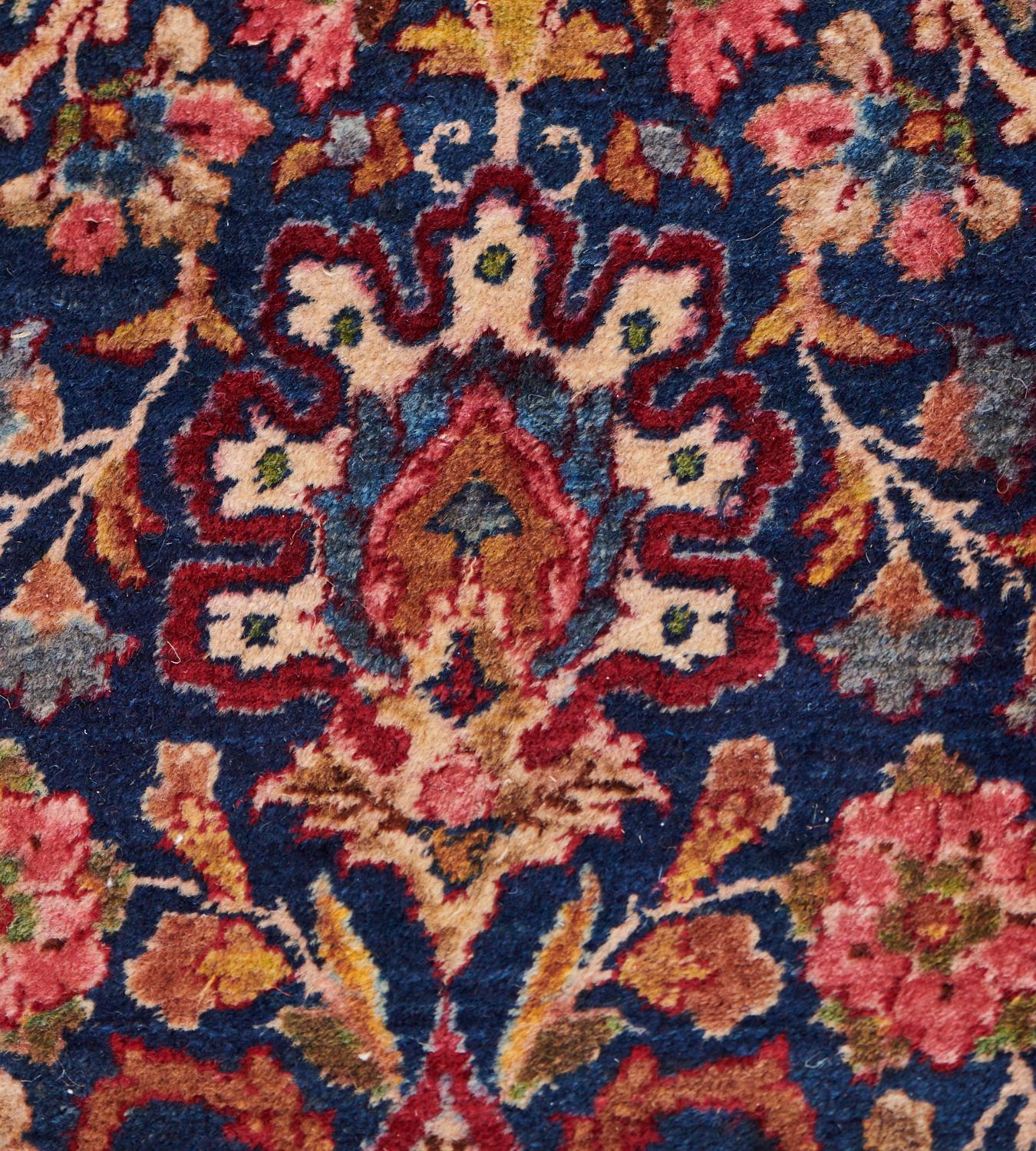 This antique, circa 1920, Kirman rug has a deep indigo-blue field with an overall design of horizontal bands of ivory cusped palmettes issuing smaller palmettes containing a scrolling floral vine, surrounded by a dense polychrome floral vine linking