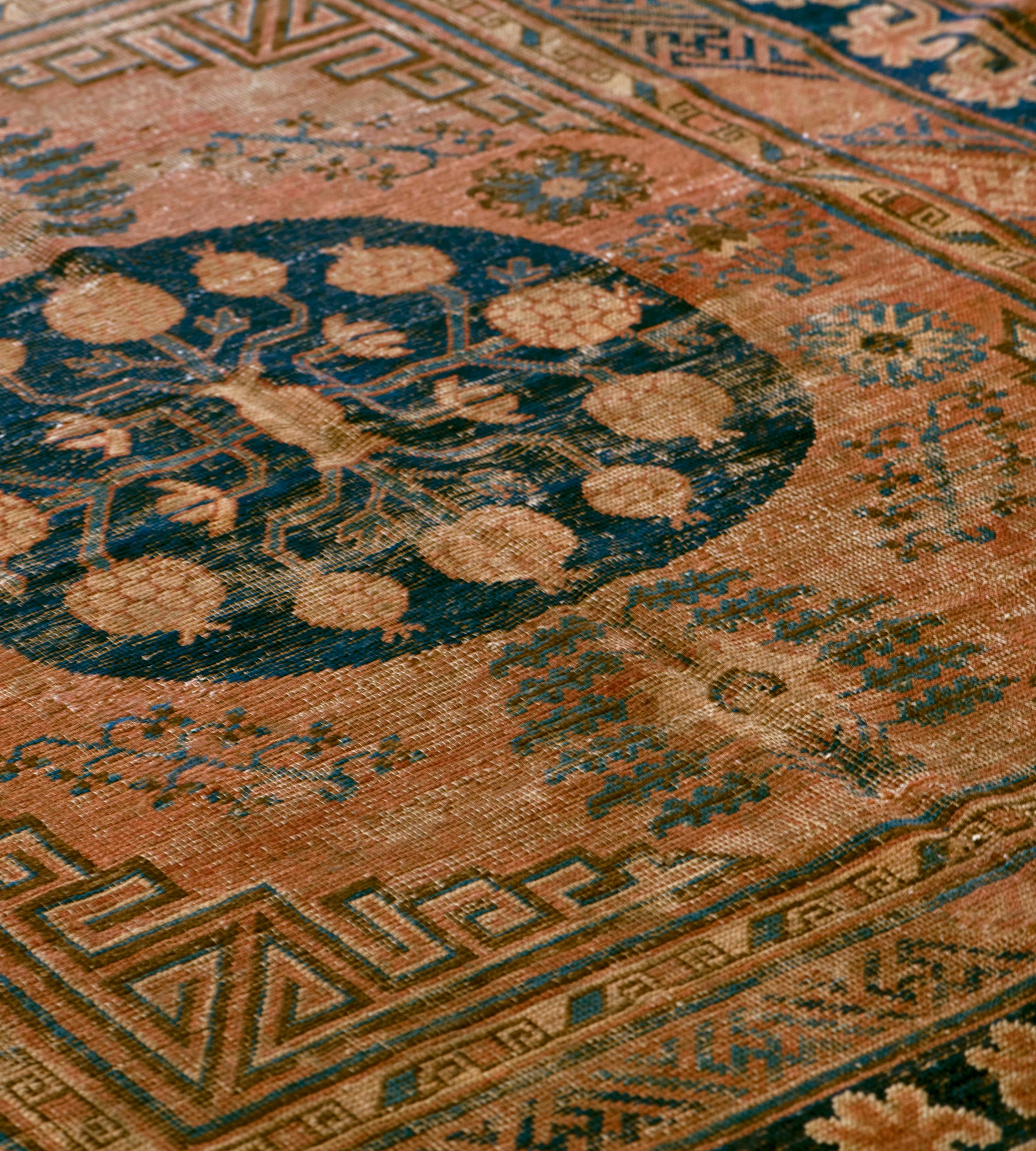 This traditional Khotan rug has a shaded terracotta-red field scattered with a variety of floral sprays, hooked lozenges and palmettes radiating angular leaves around a royal-blue roundel with a central buff-brown vase issuing angular vine with a
