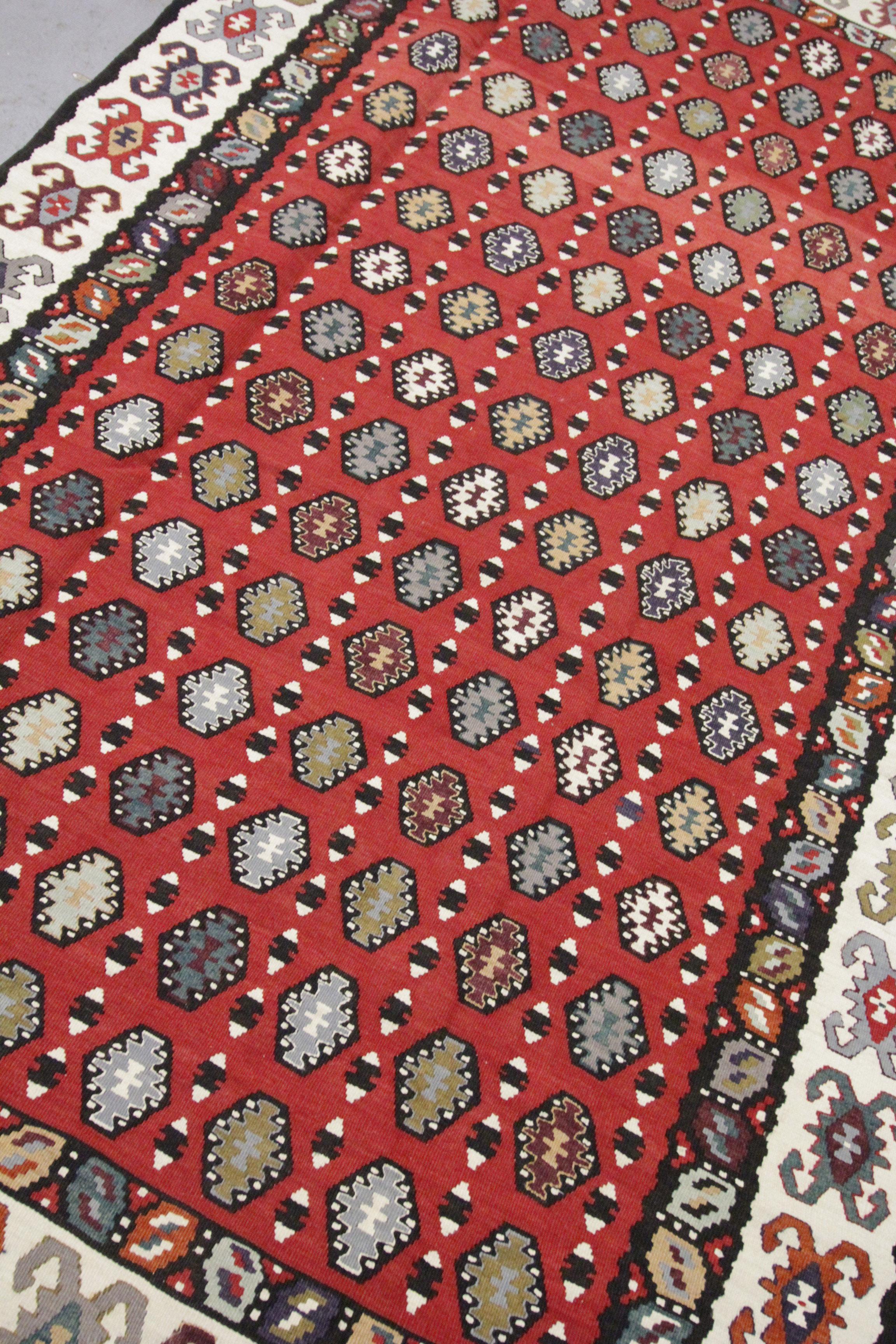 This beautifully woven bold rug is a handwoven kilim that was incorporated in the early 20th century. The design is bold with a rich red background and repeating motifs woven in blue, grey and mustard accents. A repeating pattern border then