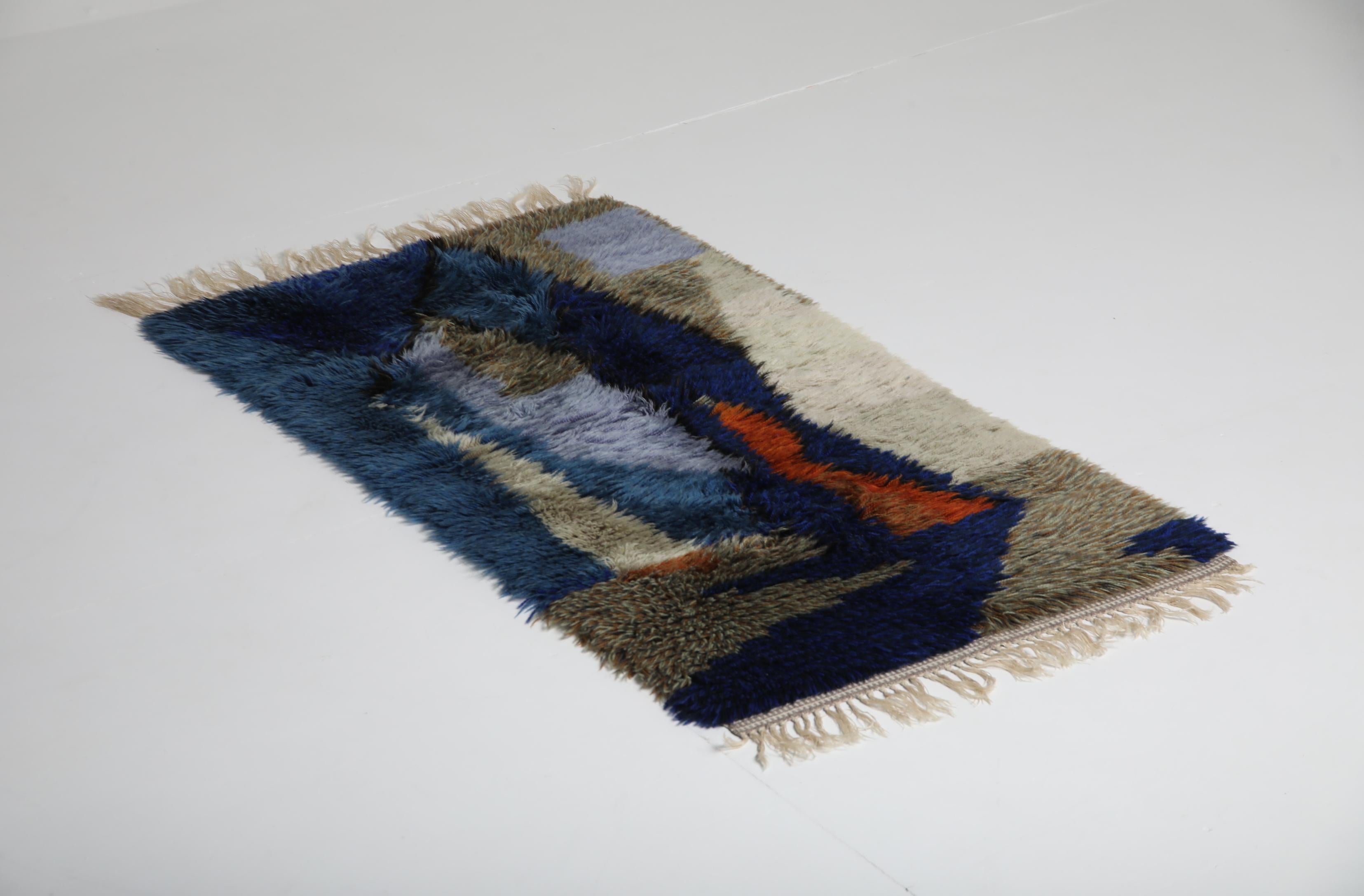Alfombra escandinava rya tejida a mano en la década de 1970, caracterizada por su pelo profundo y su composición geométrica en tonos estratificados de azul, beige, gris y naranja quemado. La alfombra está realizada en pura lana, lo que le confiere