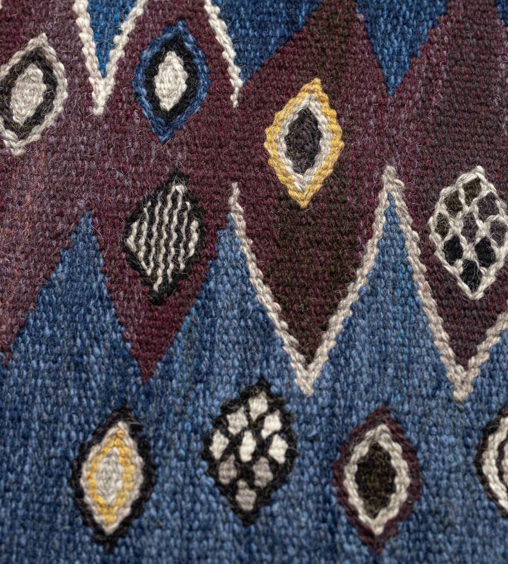 This signed mid-century Swedish rug has a shaded blue field with three central columns of linked chocolate-brown and soft-brown sharp lozenges outlined in ivory, flanked at each side by chocolate-brown part-lozenges, diagonal bands of polychrome