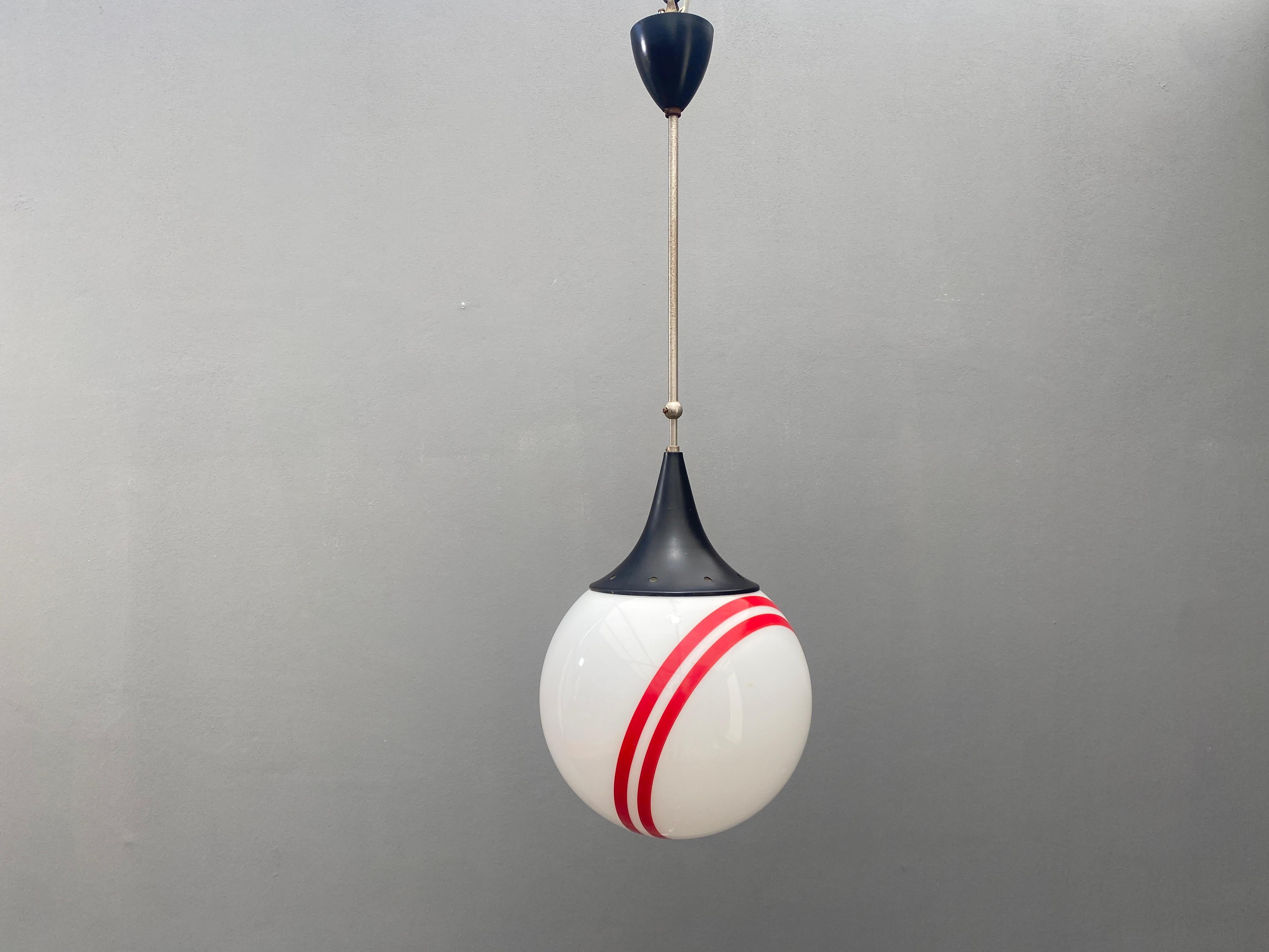 Lampe suspendue avec un abat-jour rond en verre blanc à rayures rouges. L'abat-jour est fixé à une structure métallique avec des éléments en plastique noir. Avec son design simple et ses accents de couleur frappants, il s'adapte à divers intérieurs.