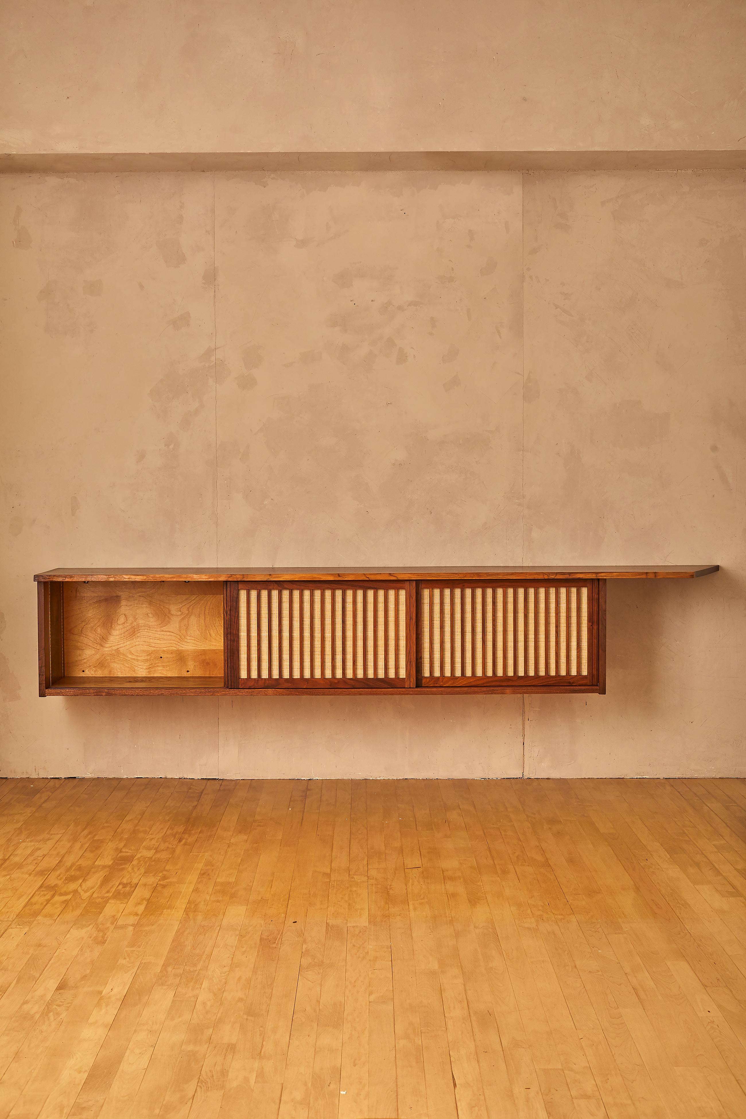 Hanging wall cabinet by George Nakashima featuring a single board plank top with free-edge, elongated expressive detail atop case with three sliding doors with grass-cloth concealing three shelves and four drawers.