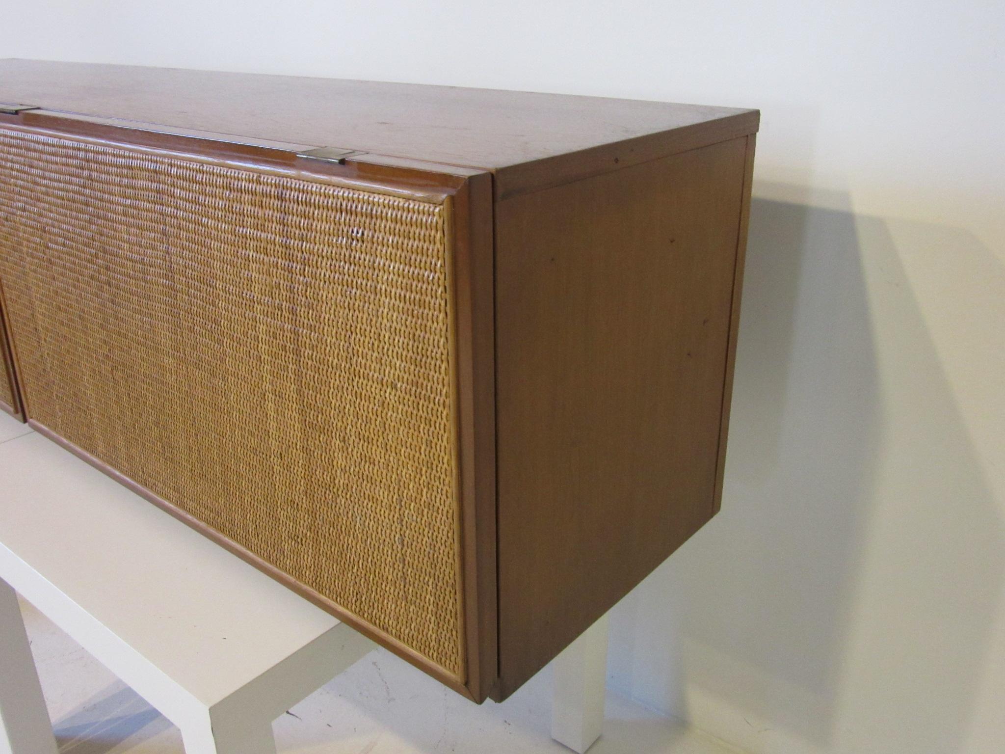 A custom ordered walnut hanging cabinet with two woven cane doors on brass hinges that flip up and rest on the top of the piece revealing double storage banks. From a National Historic Landmark Building built in 1954 in Columbus Indiana the mid