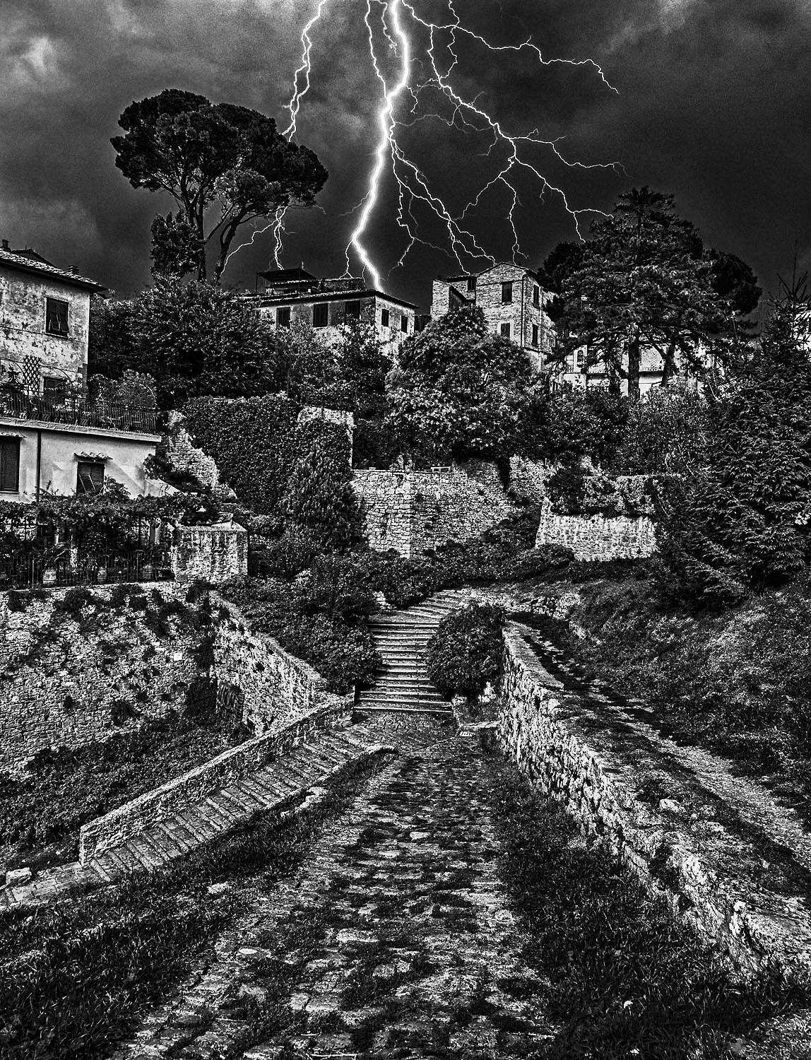 Storm, Volterra, Italy, 2015 von Hank Gans ist eine 24" x 18" große Schwarz-Weiß-Landschaftsfotografie, die einen Sturm in Volterra, einer ummauerten Stadt südwestlich von Florenz in der Toskana, festhält. 

Dreieckige Formen bestimmen das Foto,