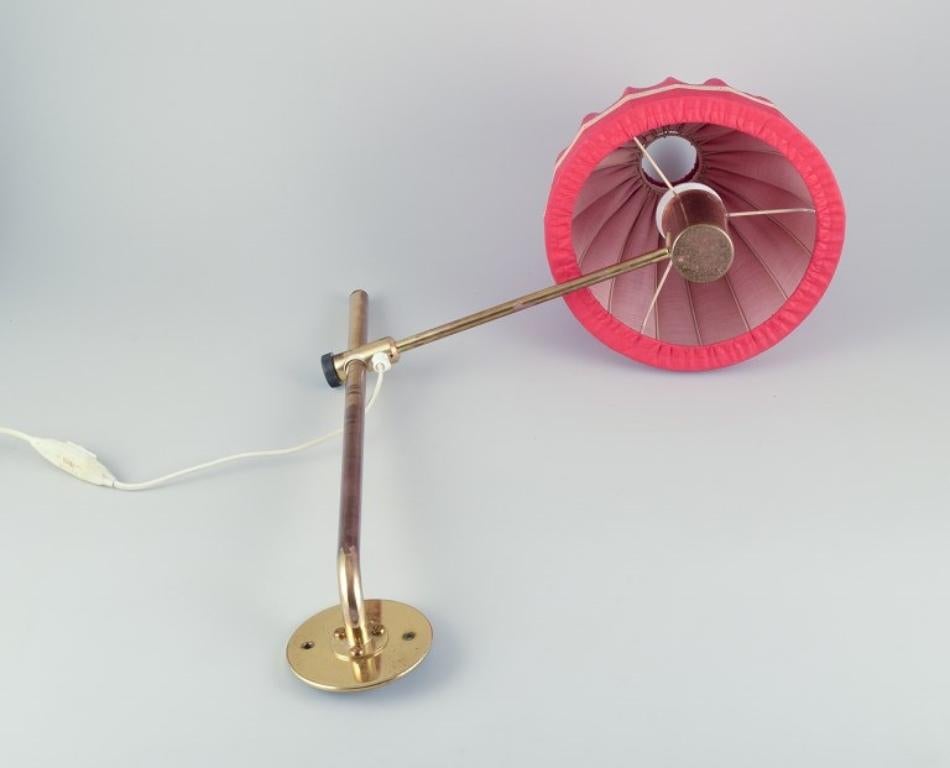 Hans Agne Jakobsson, Swedish designer. 
Wall lamp in brass with a lampshade in red fabric. Modernist design.
Circa 1960s/70s.
Label.
Max 100 watts.
In excellent condition with natural wear.
Dimensions: H approx. 55.0 cm including shade x W approx.