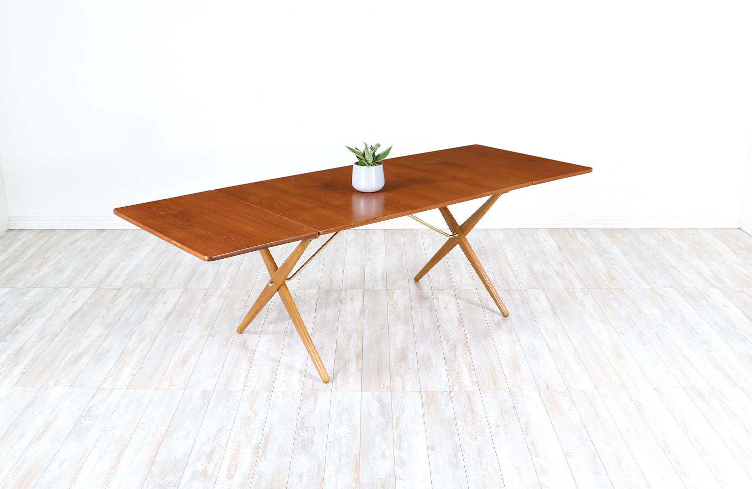 X-legged dining table designed by Hans J. Wegner for Andreas Tuck in Denmark in 1950s. The iconic AT-309 table is comprised of a teak wood top with two drop-leaves. The crossed oak wood X legs are supported by brass metal struts that provide