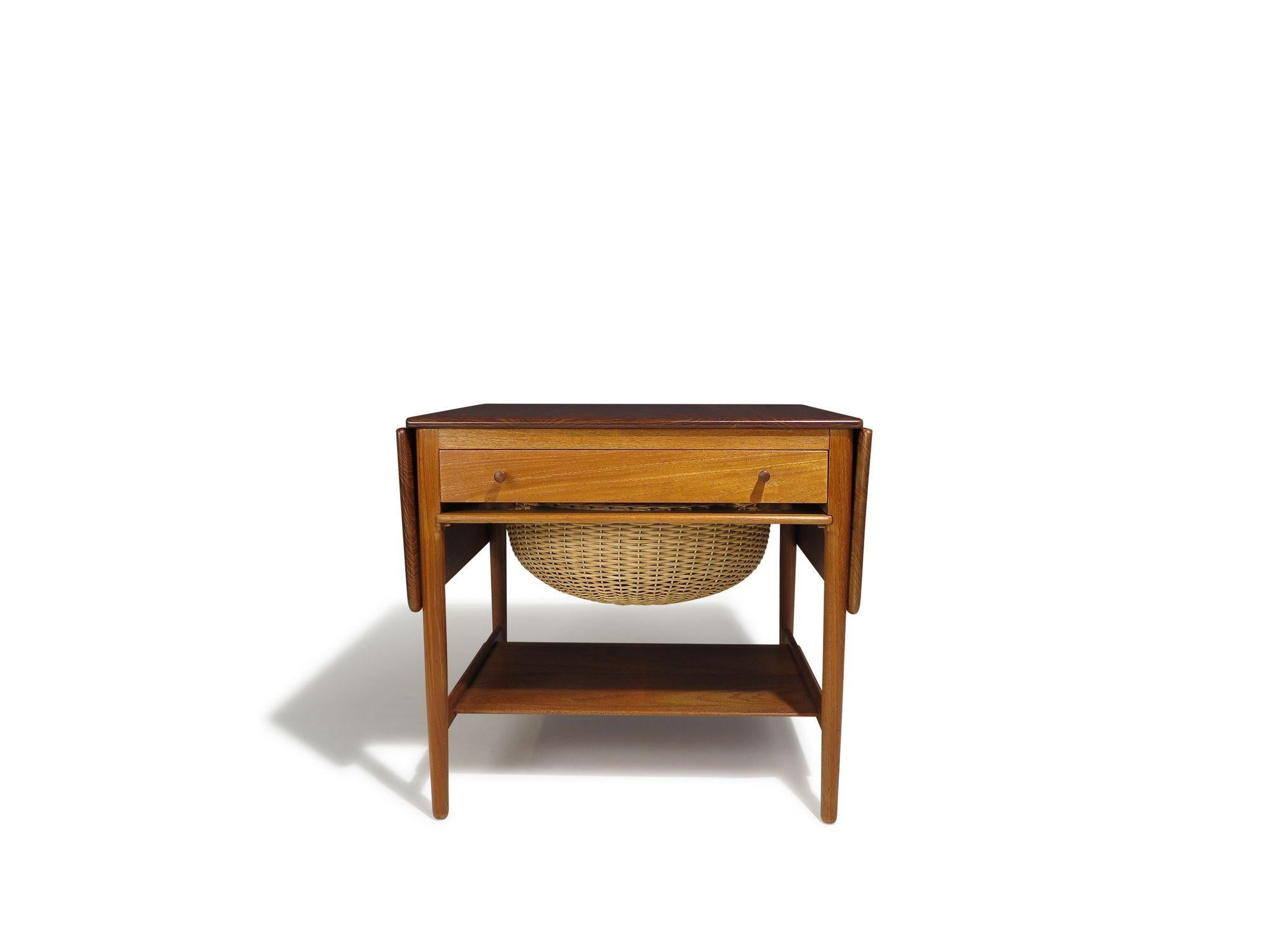 Danish teak side table by Hans J. Wegner for Andreas Tuck, Model AT-33 . The table crafted of solid teak features two drop leaves, a drawer with built in compartments for sewing materials, a pull-out wicker basket for additional storage, and a lower