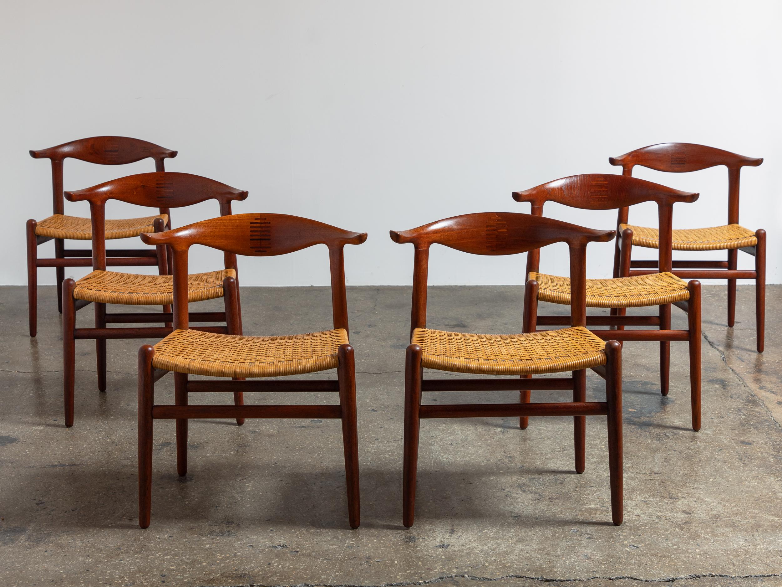 Un set coordinato di sei sedie JH-505 Cowhorn, disegnate da Hans J. Wegner per Hansen. Realizzate in teak con sedili in canna, queste sedie iconiche si distinguono per i loro braccioli elegantemente curvati, che ricordano le corna di una mucca,