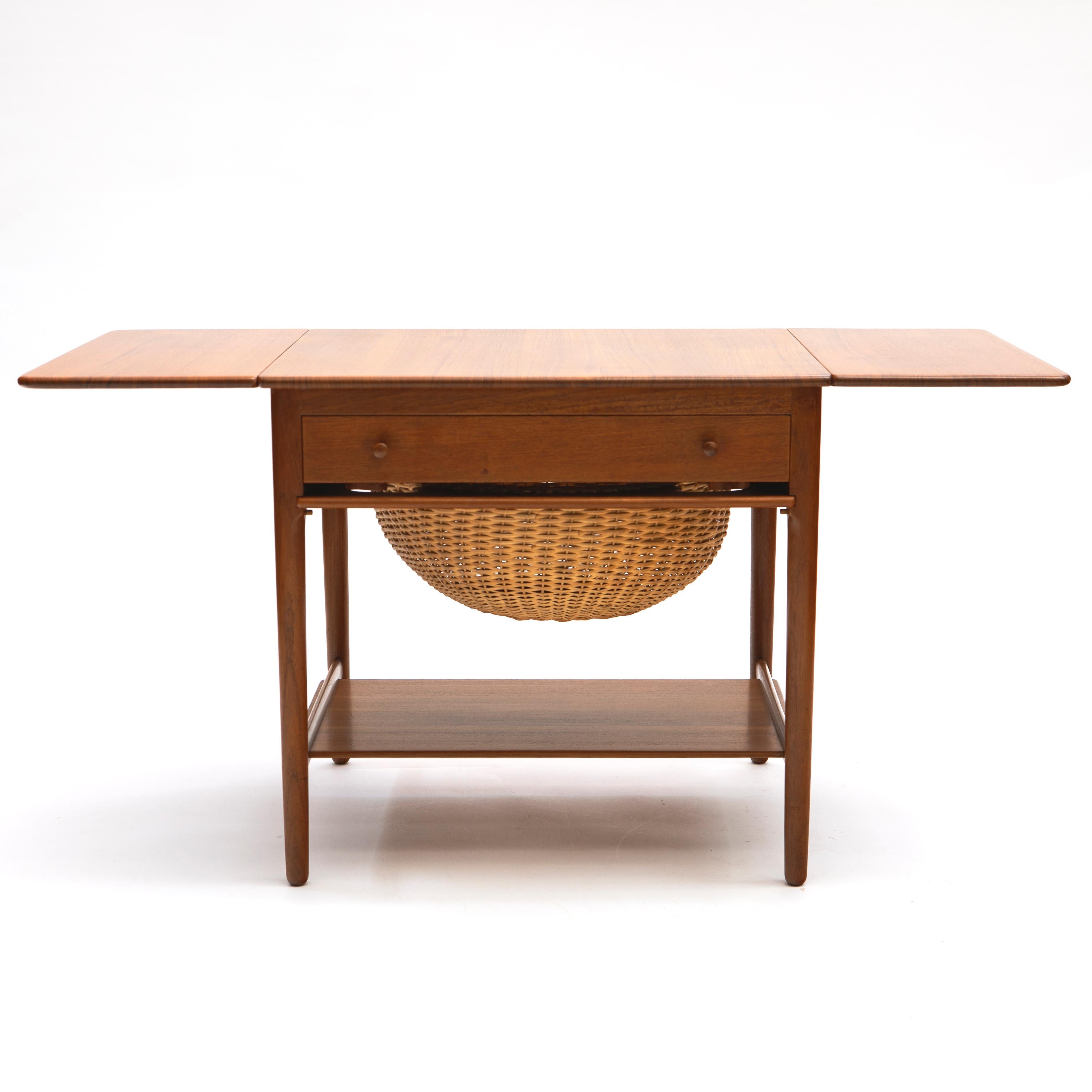 Sewing table, model AT-33 in solid teak.
Features drop leaves, a wicker basket for knitting’s, and a drawer with spindles and divisions for thread and other supplies. 
Designed by Hans J. Wegner manufactured in Denmark by cabinet maker Andreas Tuck,