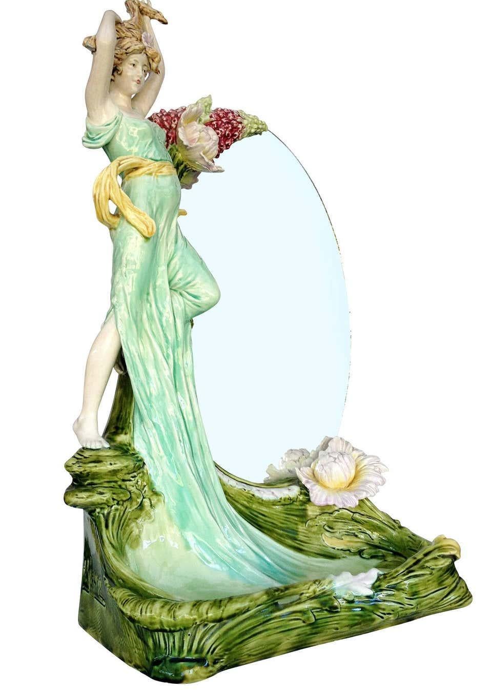 This oval vanity mirror is decorated with a large 23-inch ceramic female figure statue with Art Nouveau floral patterns. A large tray in front of the mirror is decorated with an organic leaf pattern. The tray can hold jewelry or small hand towels.