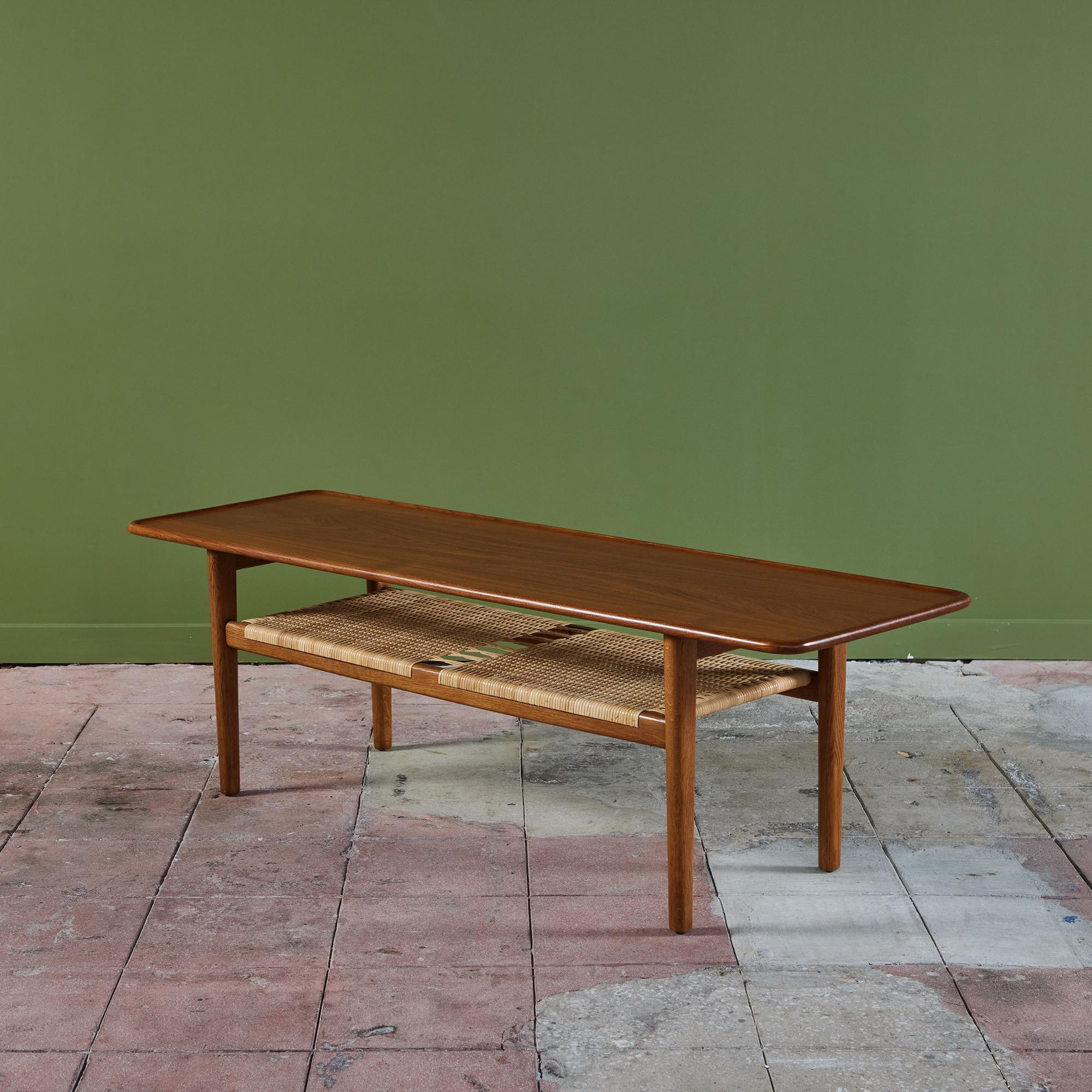 Table basse conçue par Hans Wegner pour Andreas Tuck en 1955. Un large plateau à bord arrondi et surélevé est fabriqué en teck massif et repose sur une base en chêne. L'étagère à magazines située en dessous est recouverte de canne tressée.
