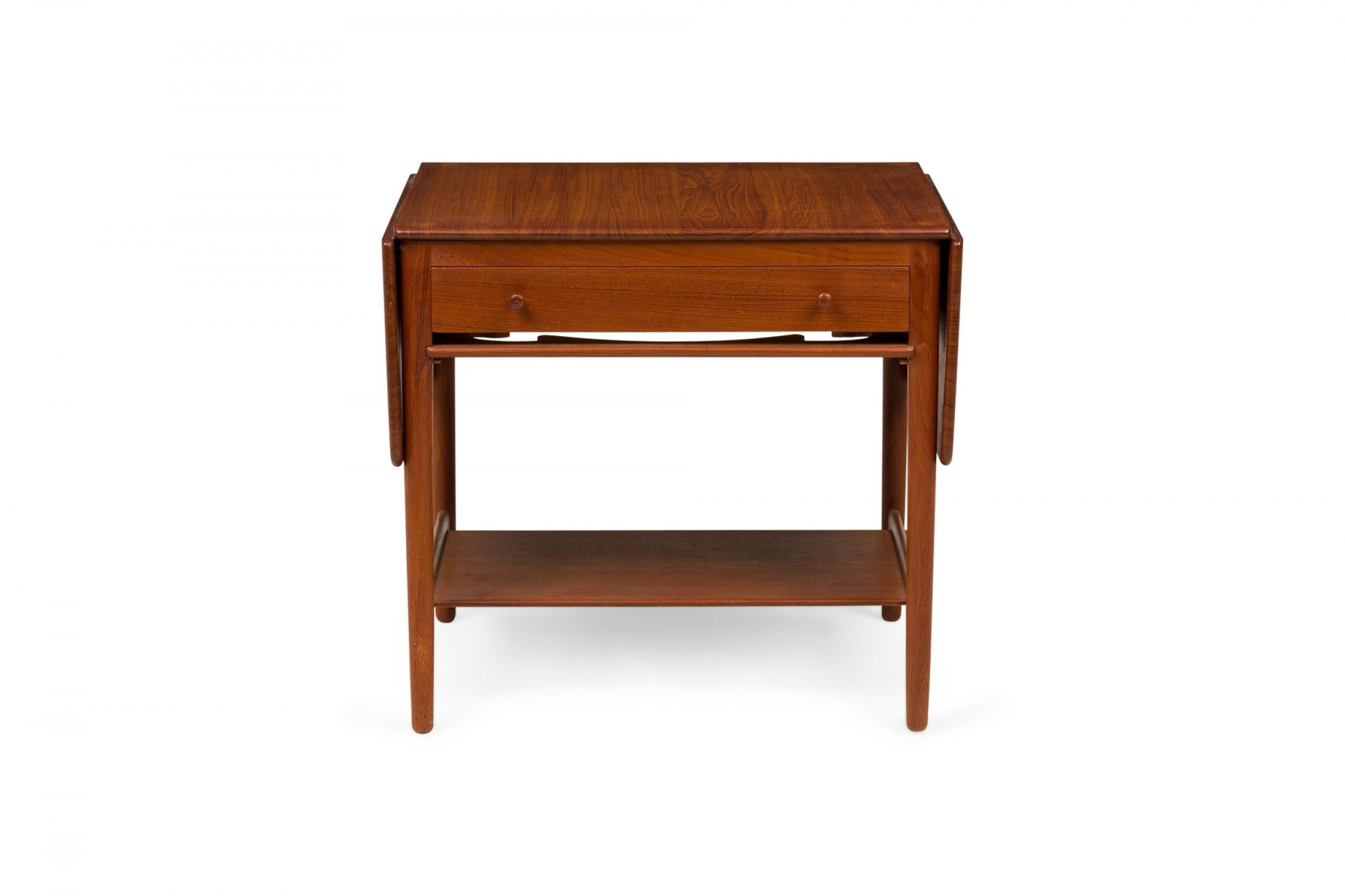American Mid-Century teak wood sewing table with two drop leaves on either side, a single drawer with two wooden knob drawer pulls, and a sliding compartment with room for a basket over a smaller stretcher shelf connecting four tapered dowel legs.