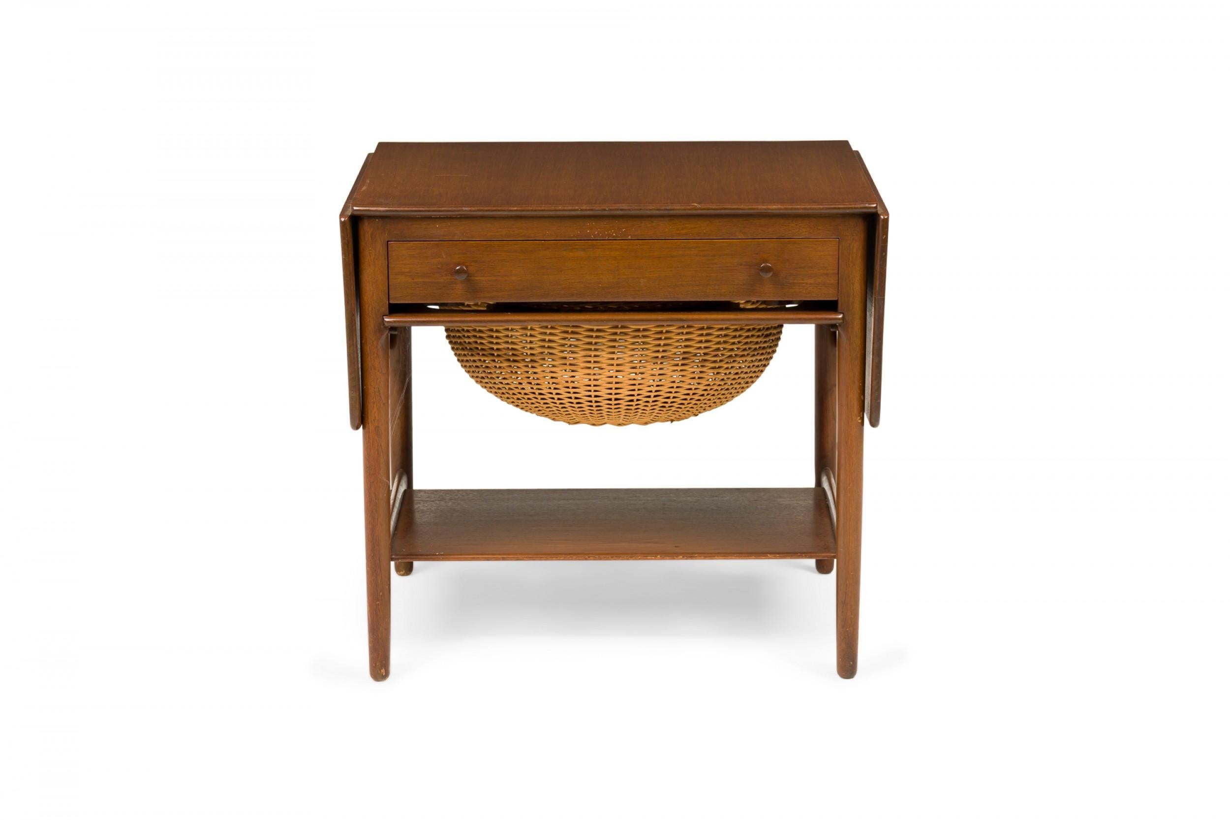 American mid-century teak wood sewing table with two drop leaves on either side, a single drawer with two wooden knob drawer pulls, and a sliding compartment containing a removable woven wicker basket over a smaller stretcher shelf connecting four