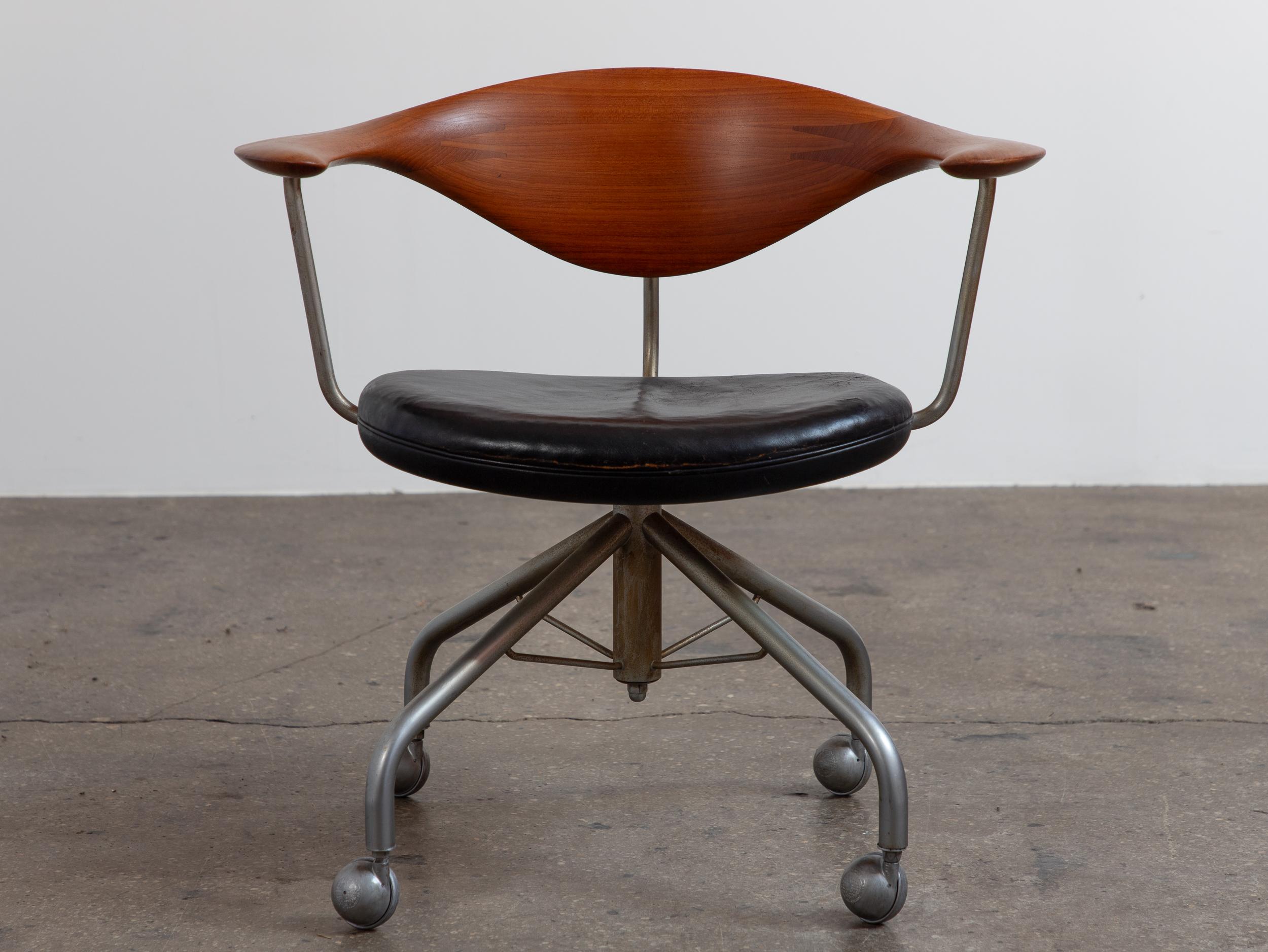 Sedia da ufficio girevole modello JH502, disegnata da Hans Wegner per Hansen. Lo schienale e i braccioli scultorei in teak presentano fantastiche venature del legno e le iconiche giunture a pettine del designer. La seduta scultorea offre comfort e