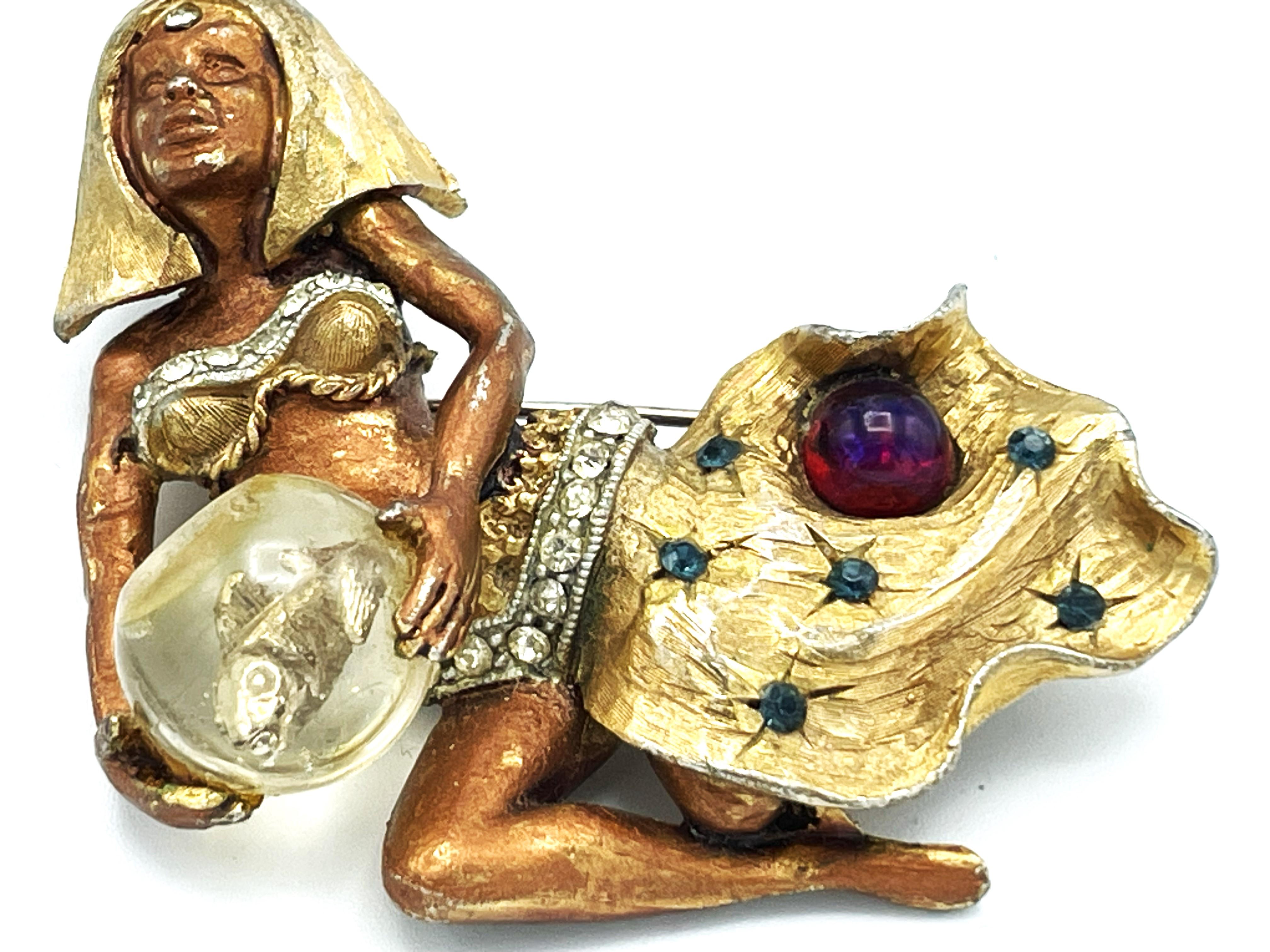 This fantastic HAR brooch is a rare find! The gold toned Cleopatra holds a bowl with a gold-toned metal fish inside. Her top and belt are set with clear rhinestones. The skirt features dark blue rhinestones and a single, dark red ball with a