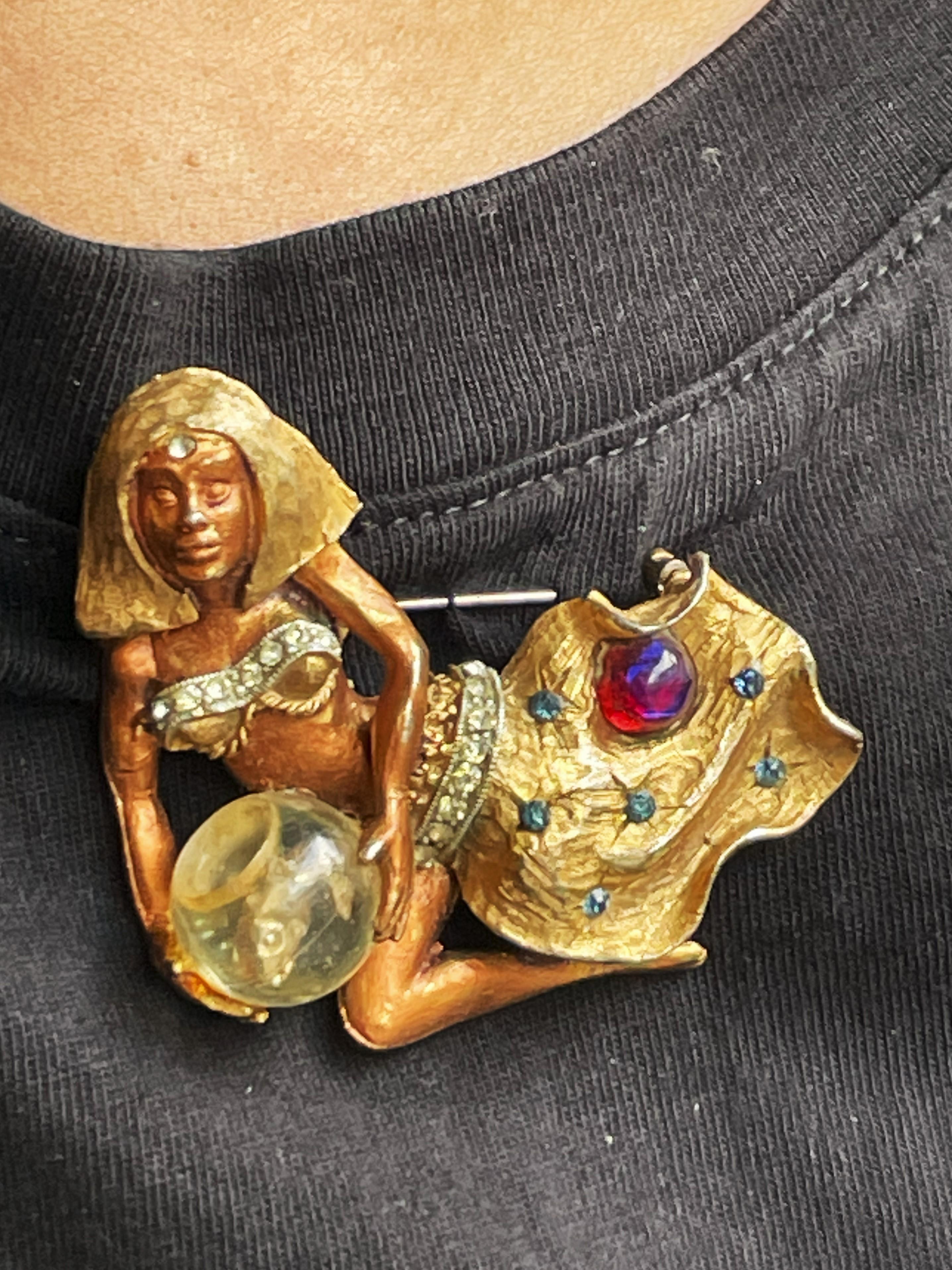 Questa fantastica spilla di HAR è un reperto raro! La Cleopatra dorata tiene in mano una ciotola con all'interno un pesce in metallo dorato. Il top e la cintura sono impreziositi da strass trasparenti. La gonna presenta strass blu scuro e un'unica
