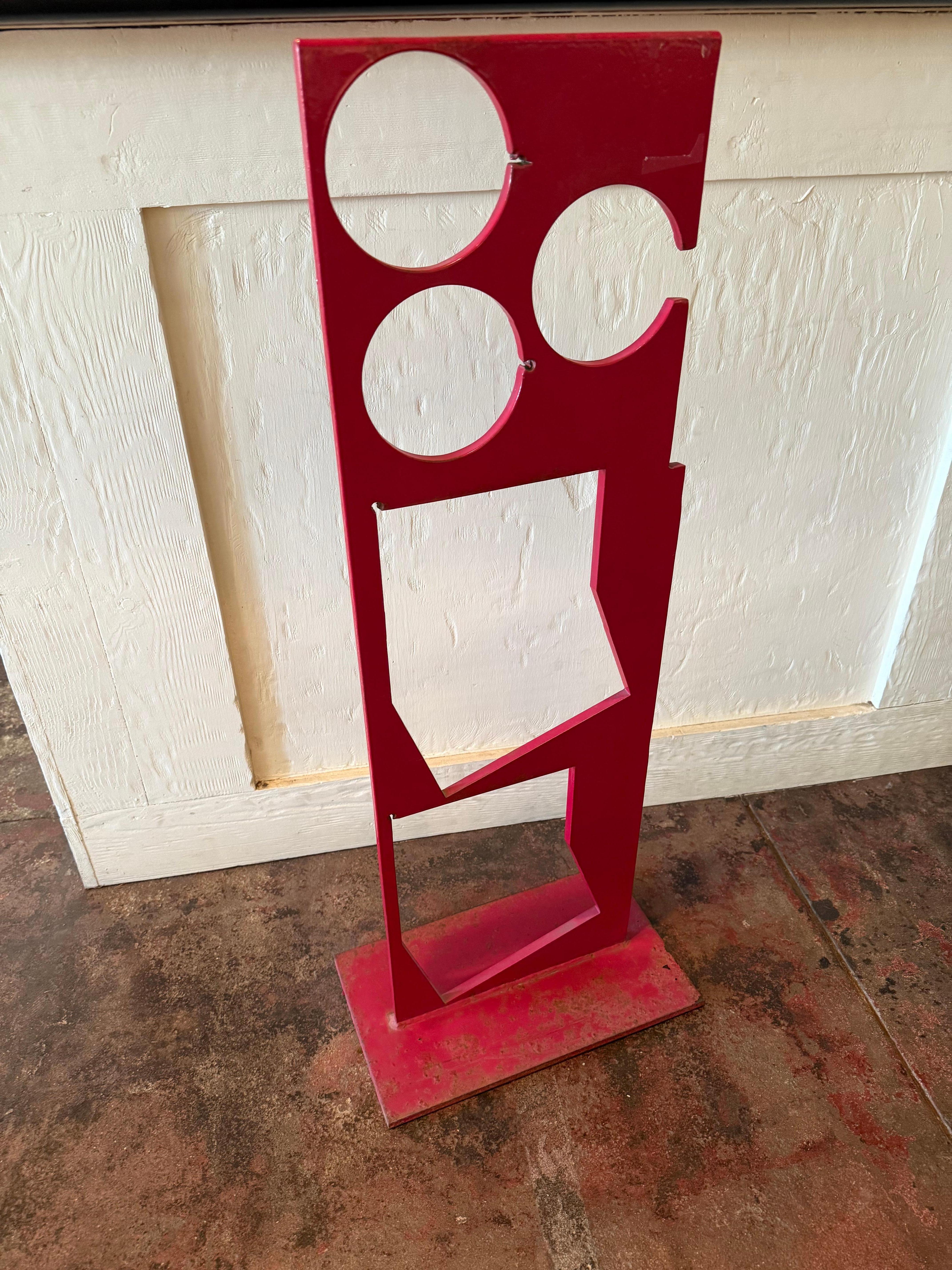 A freestanding hard-edge sculpture in vivid red-painted metal, composed of layered circular and angular cutouts. 
It feature simple, geometric shapes  squares, rectangles and circles, arranged in precise and symmetrical compositions. 
Edges are