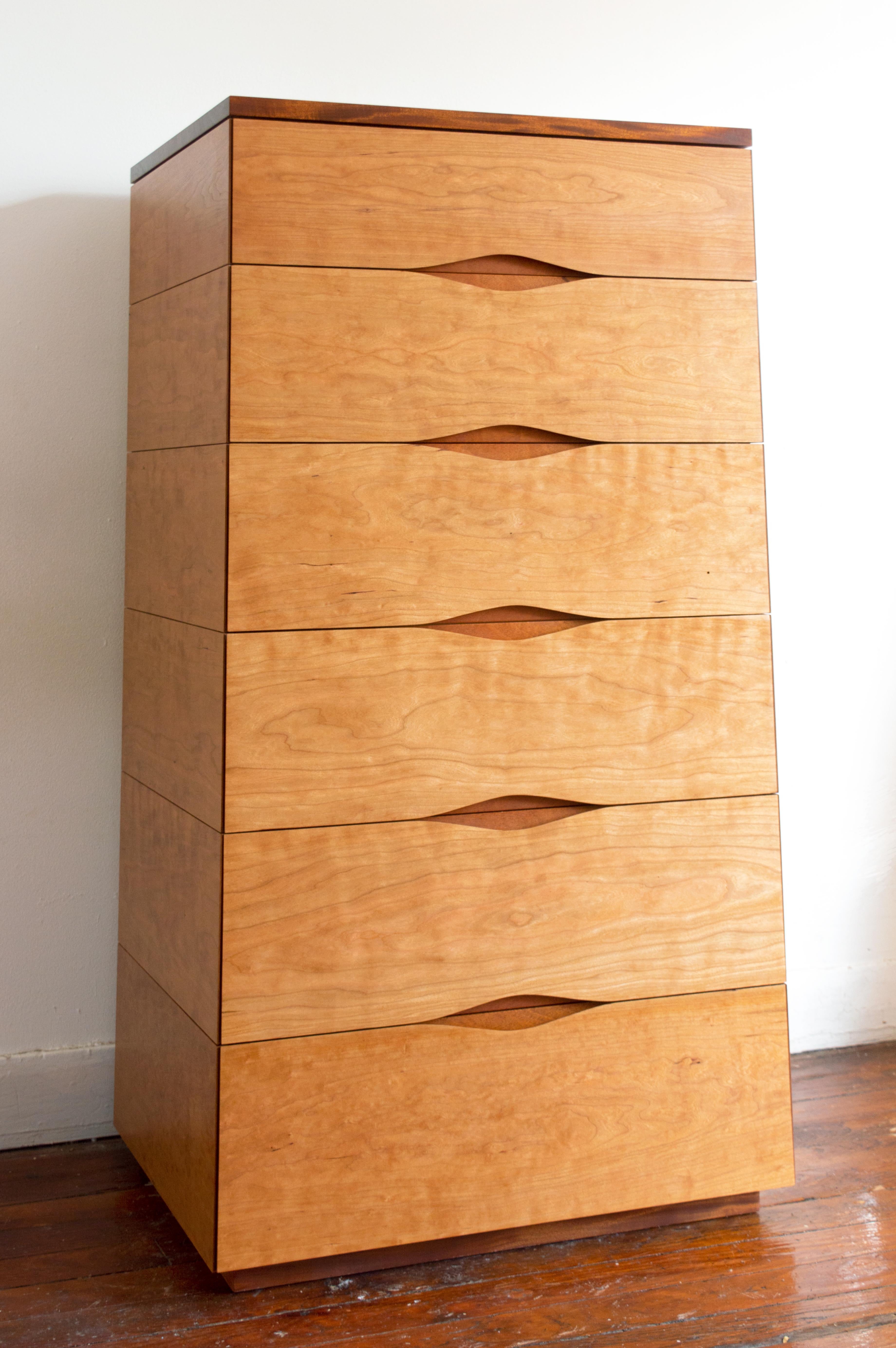 A playful monolith. The austere geometry and delineation of this tall chest of drawers are undermined by the rippling figure of the wraparound cherry cladding and the “flame” pattern of the mahogany top. 
And then there is the subtly sensuous