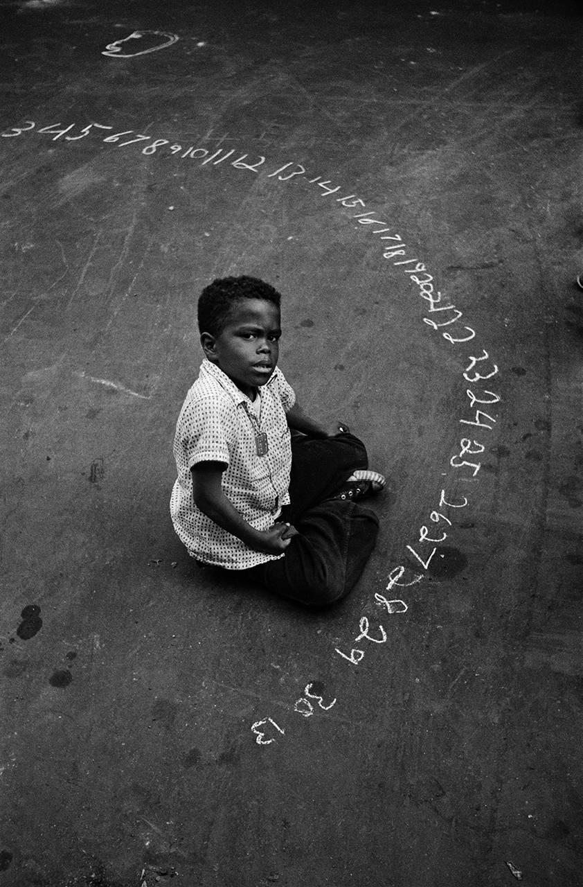 Boy with Chalked Numbers, NYC par Harold Feinstein est un tirage argentique à la gélatine de 14 x 11 pouces, et est une édition 3/200. Cette photographie représente un garçon assis par terre, entouré de chiffres écrits à la craie sur le sol. Cette