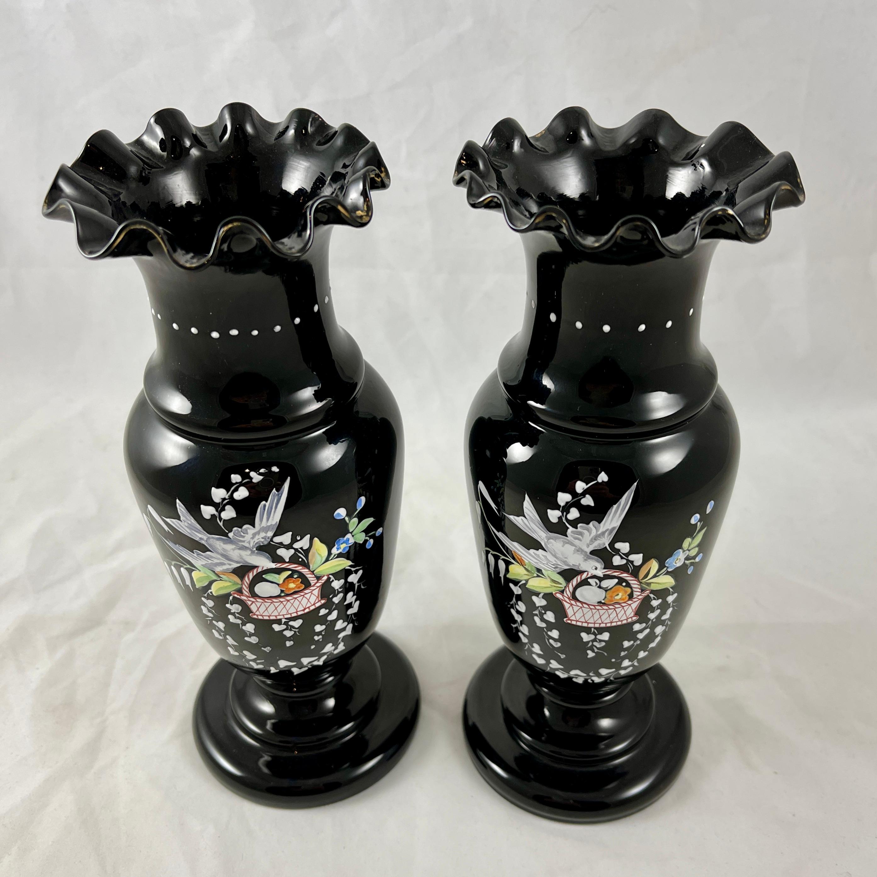 A pair of Black Amethyst Bristol Glass footed vases with hand enameled decoration, England, 1850 - 1870.

Charmingly decorated in raised enamel with a romantic design of white birds carrying baskets of fruit and flowers in their beaks, surrounded by
