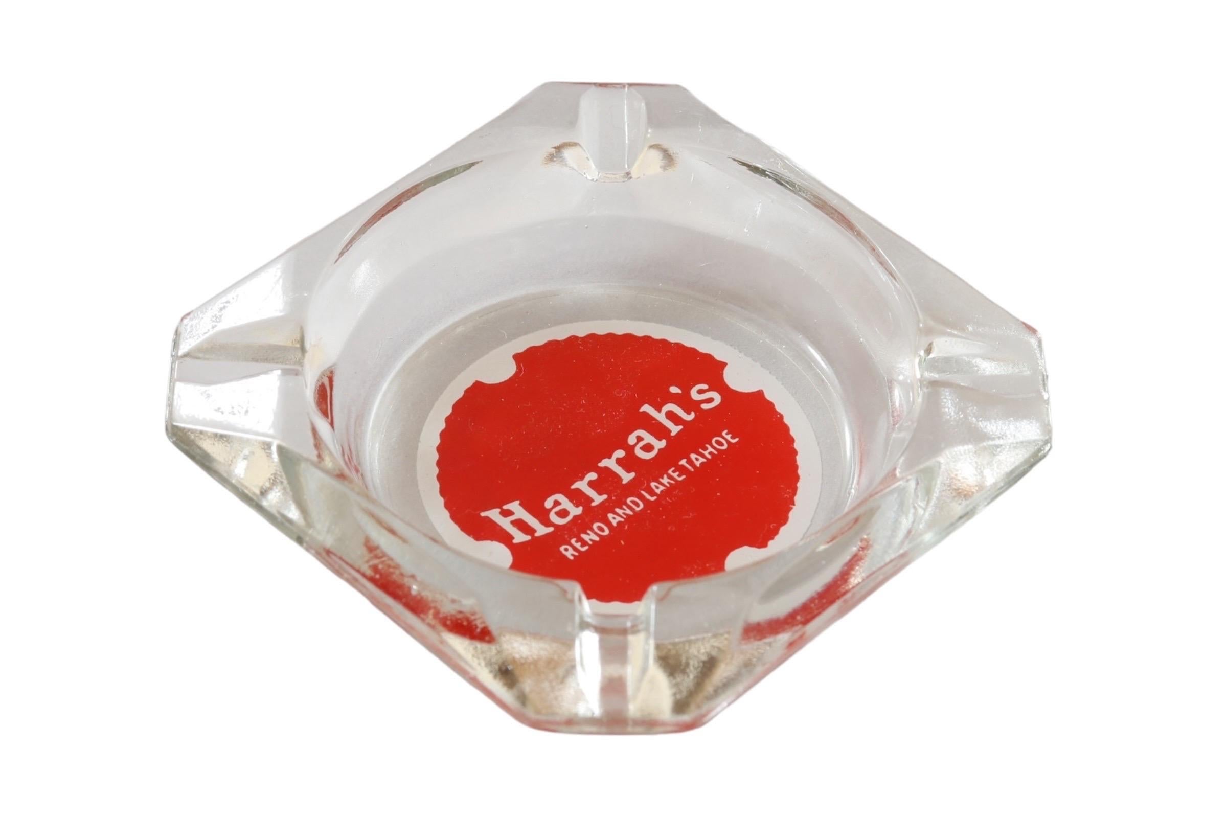 A square clear glass ashtray from the Harrah’s Hotel of Reno and Lake Tahoe, Nevada. Corners are cut, each with a cigarette rest. The center is printed with a red over white circle branded with their logo. Founded by William F. Harrah (1937–1978),