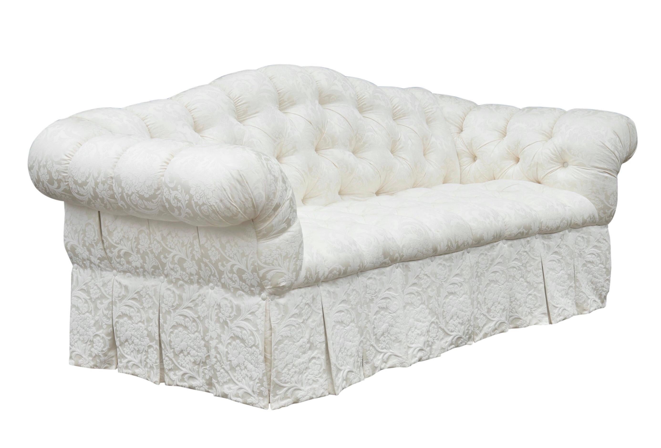 A traditional tufted camel back sofa made by Harrod’s of London. Upholstered in an elegant ivory damask with diamond button tufting and pleated rolled arms. The front is finished with a box pleated skirt and the back and sides are decorated with