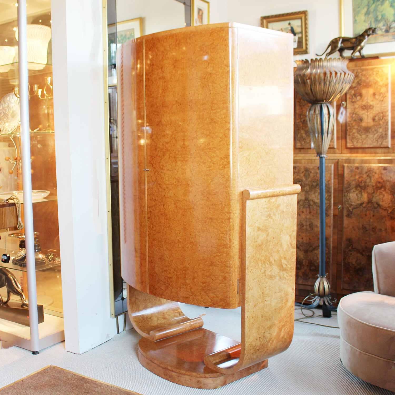 An Art Deco bow fronted cocktail cabinet set over a u-shaped base with scrolled detail. Mirrored, lit interior to top with peach glass shelves and mirrored slide tray. Ivorine handles. Lower cabinet. Burr walnut with satin birch