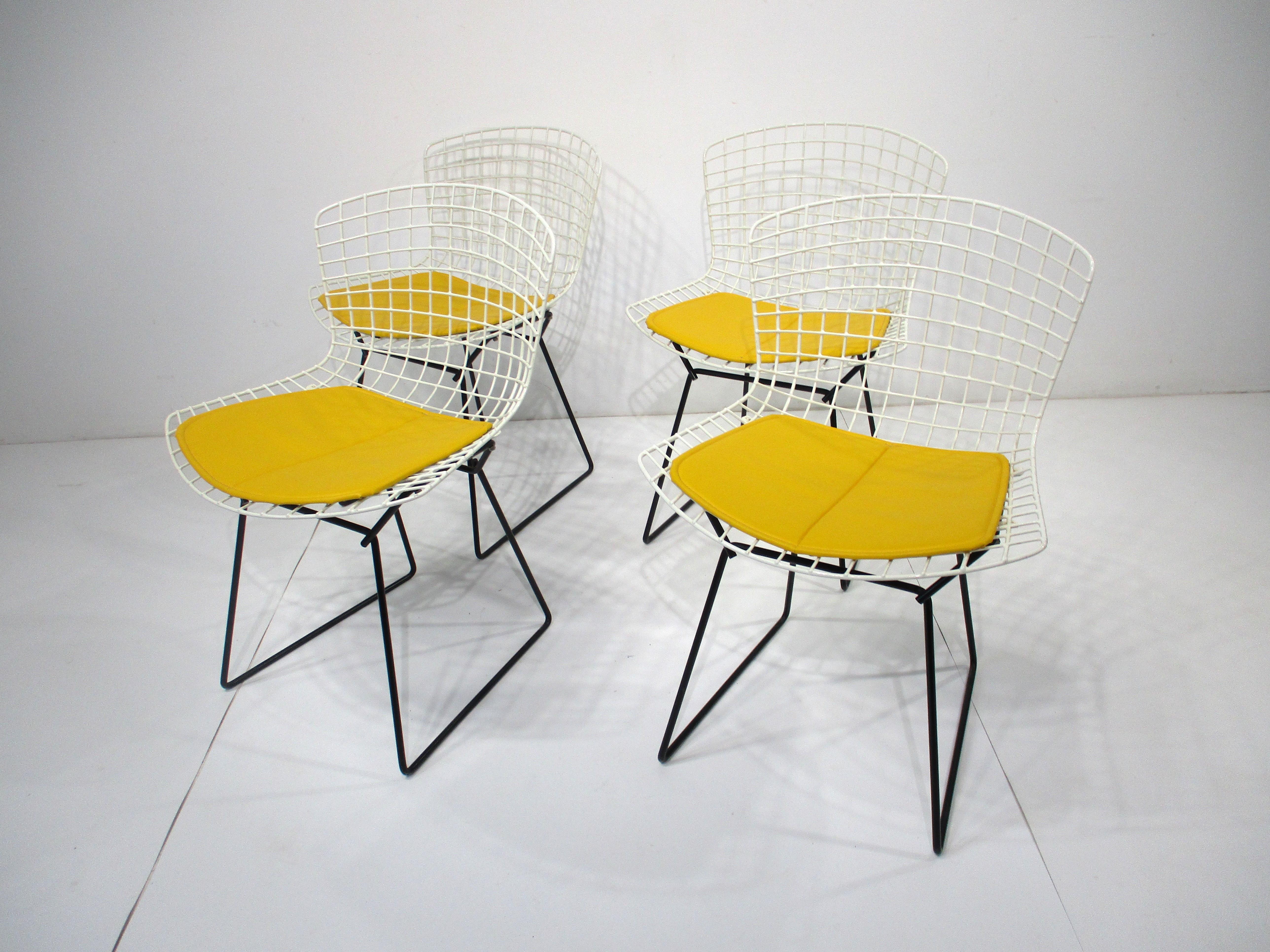 A set of four white wire dining chairs with black bases and bright yellow upholstered seat pads . A great special order combination making for a cheery and refined look and feel , designed by Harry Bertoia and manufactured by Knoll . Purchased from