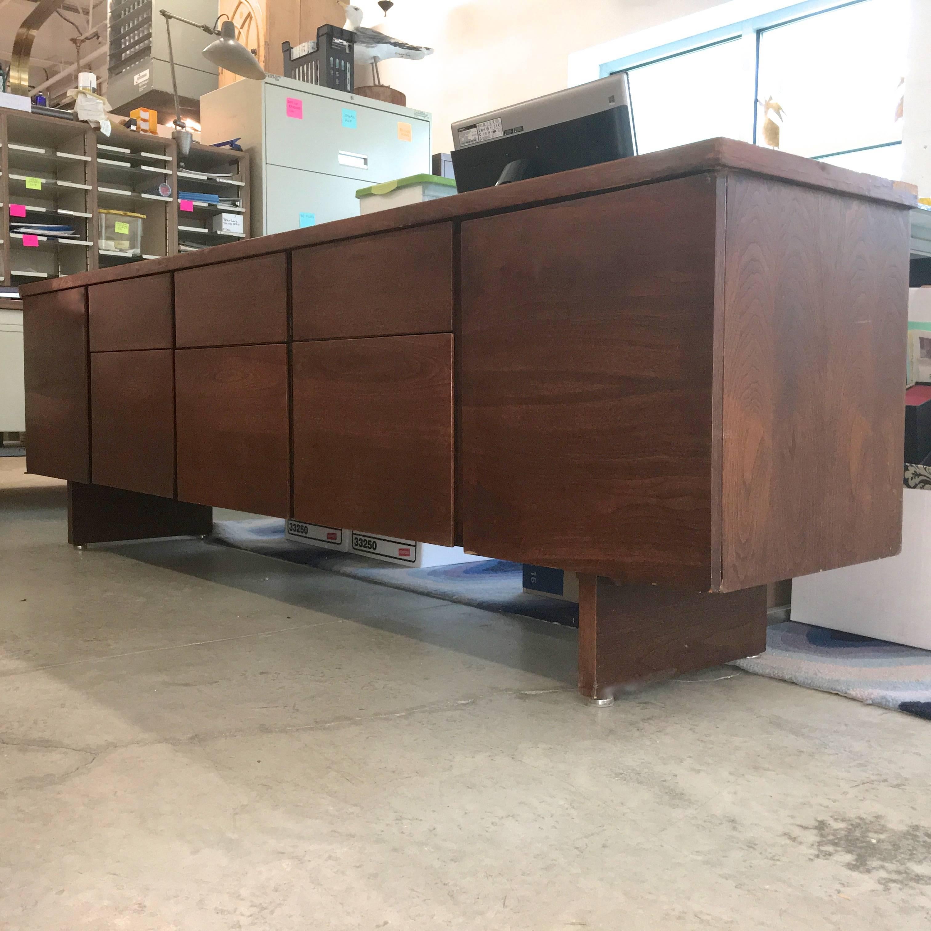 Rarely seen credenza by Harvey Probber of Fall River, MA. Long dark walnut case on two slab legs. Two doors reveal open cabinets on either end with a single adjustable shelf in each. In between the doors are three sets of drawers, shallow on top and