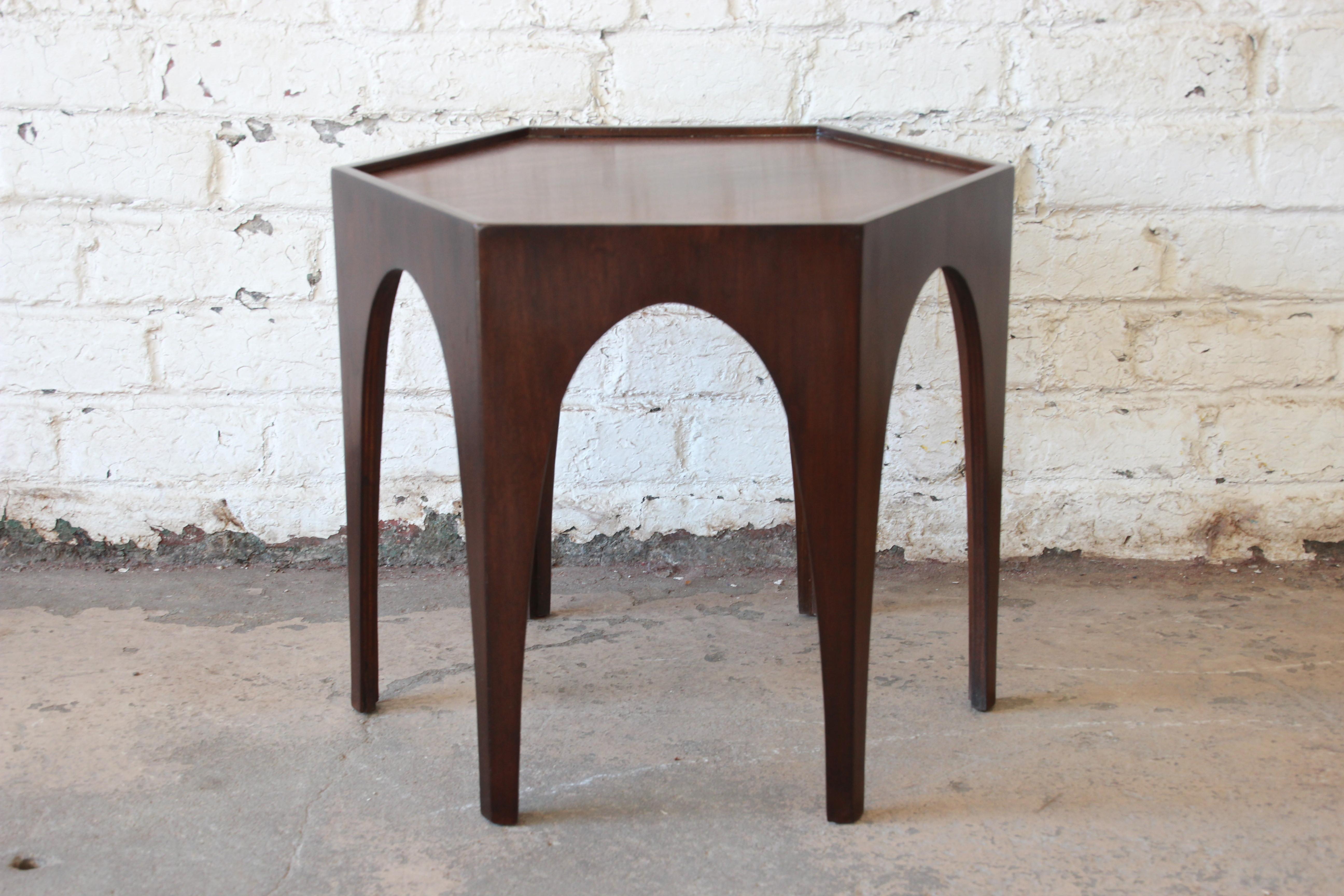 A gorgeous Mid-Century Modern hexagonal end or occasional table designed by Harvey Probber. The table features beautiful walnut wood grain and is sculpted with Moroccan-inspired arches. It is finished on all sides and could also be used as a plant