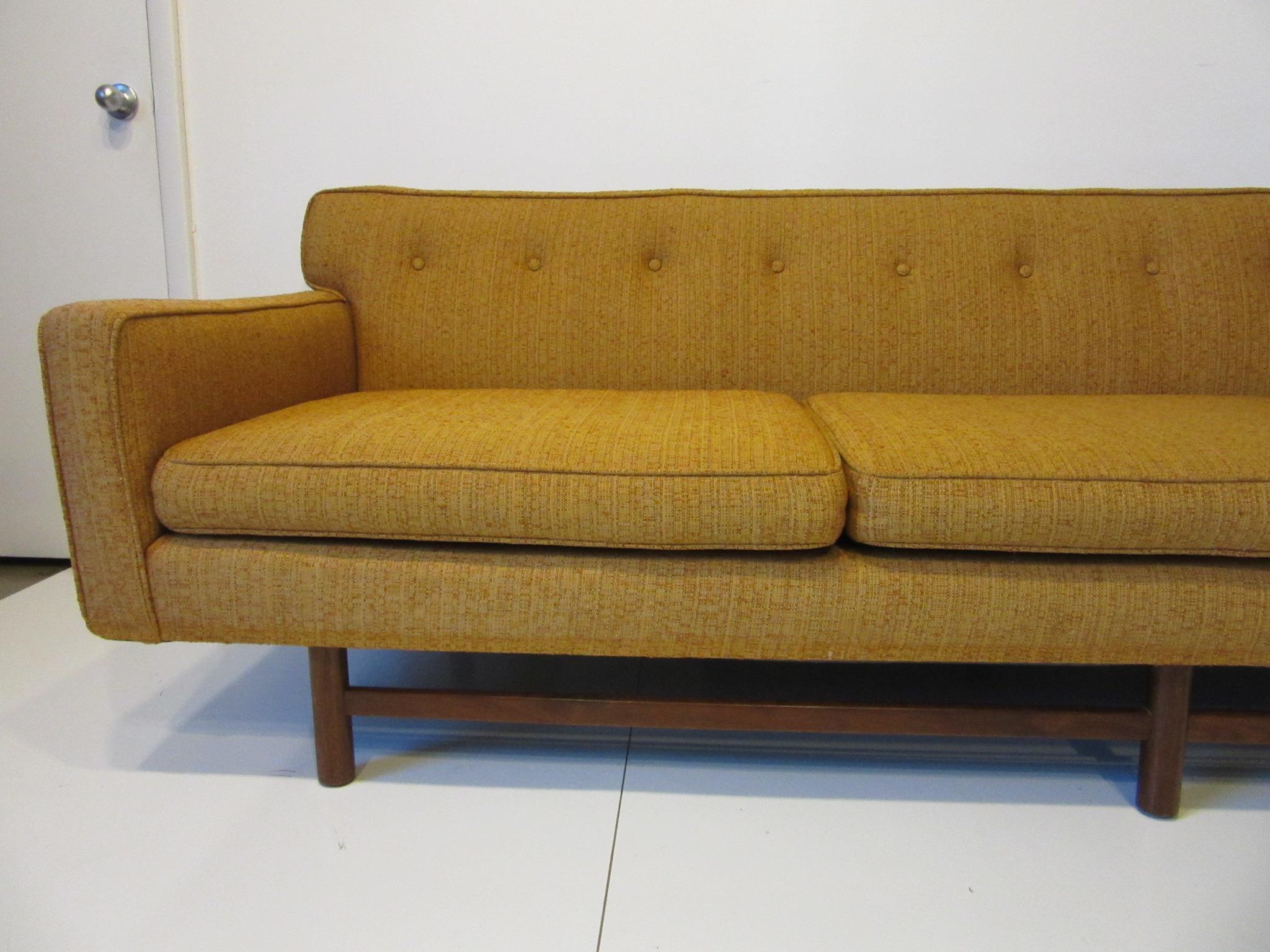 A three cushion low profile mid century sofa with button back and dark walnut legs and stretchers with overhang to each end that gives the sofa a floating affect. Designed in the manner of the Harvey Probber Furniture company, the sofa has great