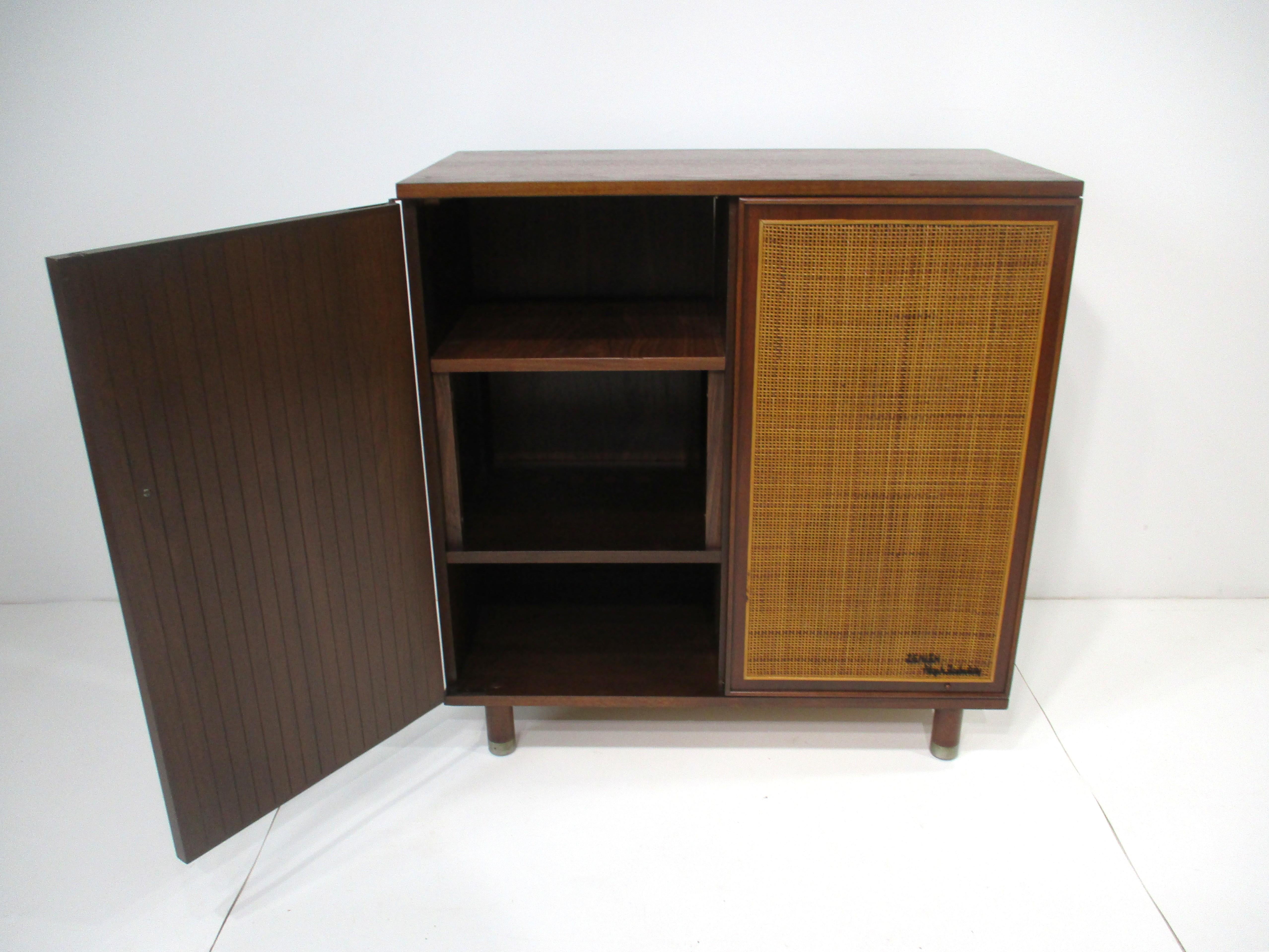 A walnut stereo record cabinet with a door having storage or for your entertainment equipment and to the other side a caned front with two speakers inside . The speakers inside seem to be one 6-8