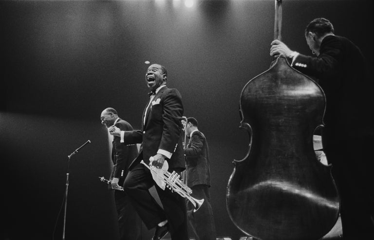 American jazz trumpeter and bandleader Louis 'Satchmo' Armstrong (1900 - 1971), shouts after clarinettist Edmund Hall's solo, on stage during the band's British tour, May 19, 1956. An unidentified bassist stands in the foreground. (Photo by Haywood
