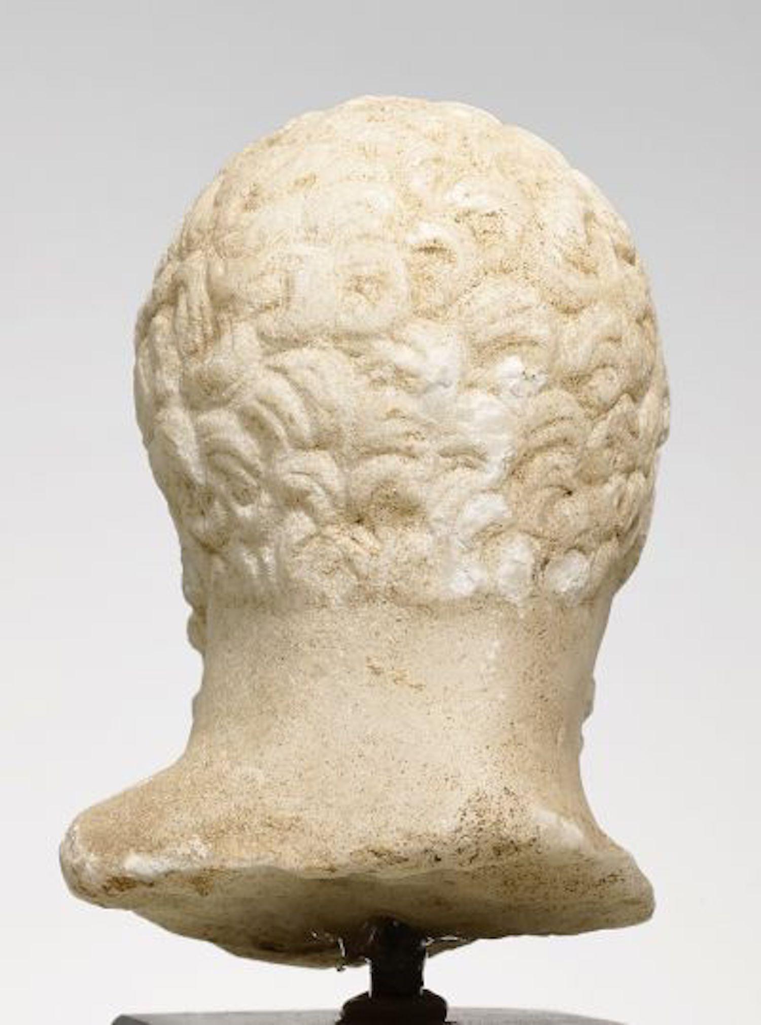 Hellenistic, ca. 2nd/1st century BC
Fine white crystalline marble.
H. 6,5 cm.

A small head of a marble figurine of the „Herakles Farnese“ type.
The hero’s head is slightly turned downwards, wearing a long wooly beard and short curly hair. A typical
