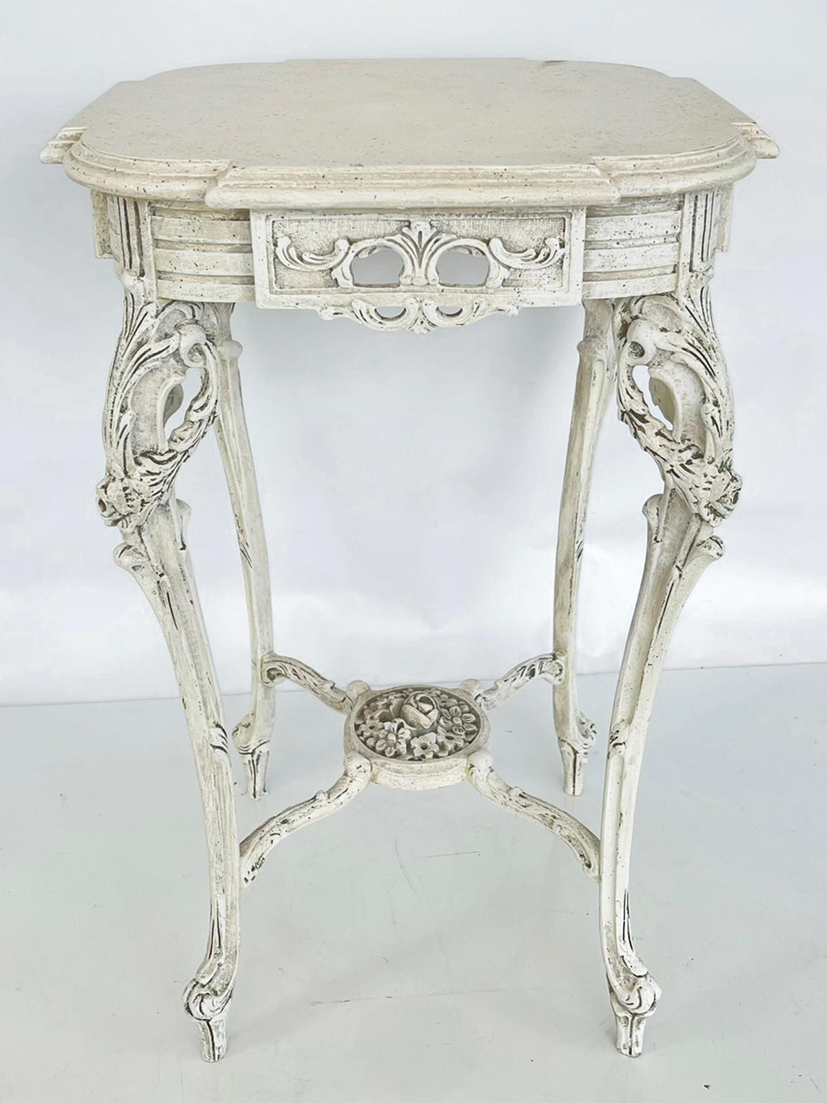 Candlestand occasional table, having an ivory-painted finish showing natural wear. Its square, molded table top with rounded corners, over a pierced, scrolling apron flanked by wide bands. The table is raised on four, acanthine-carved, cabriole