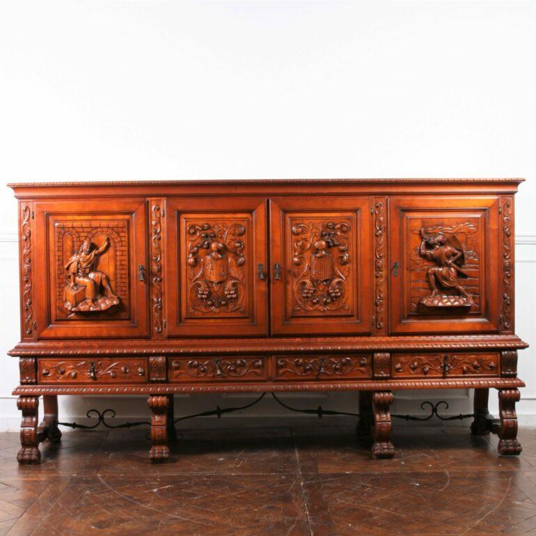 A deeply carved vintage Belgium sideboard, china or bar cabinet, of solid mahogany. Doors have three dimensional sculptures of a king, a knight, and family crests or coats of arms. Raised on solid wood legs that are joined by wrought iron braces,