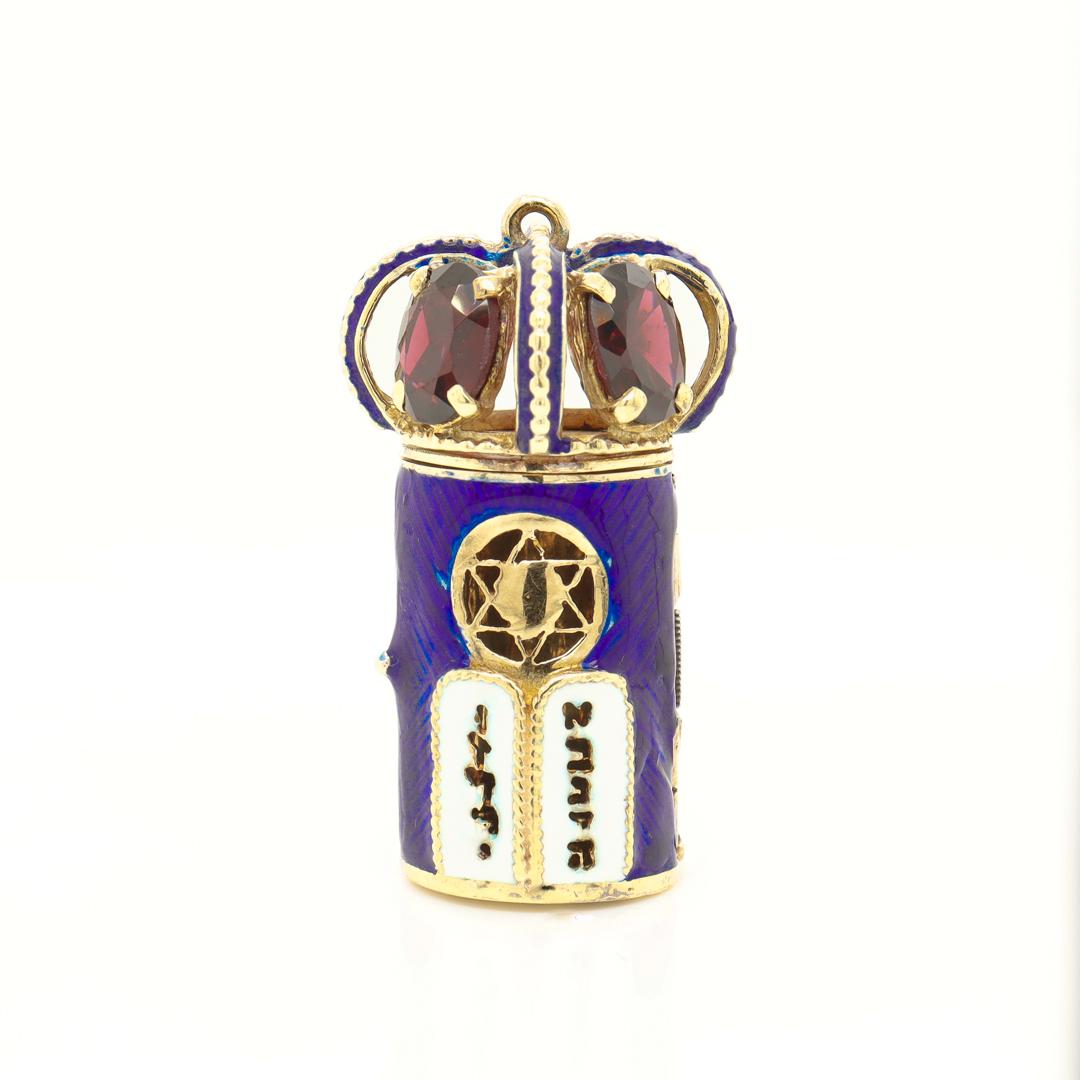 A fine Mezuzah charm or pendant.

In 14k gold with blue enamel and garnet gemstones.

In the form of a tower with a hinged door that opens, a pierced Star of David opening, and a looped crown top.

Prong-set with oval-cut garnets to the crown.

Set