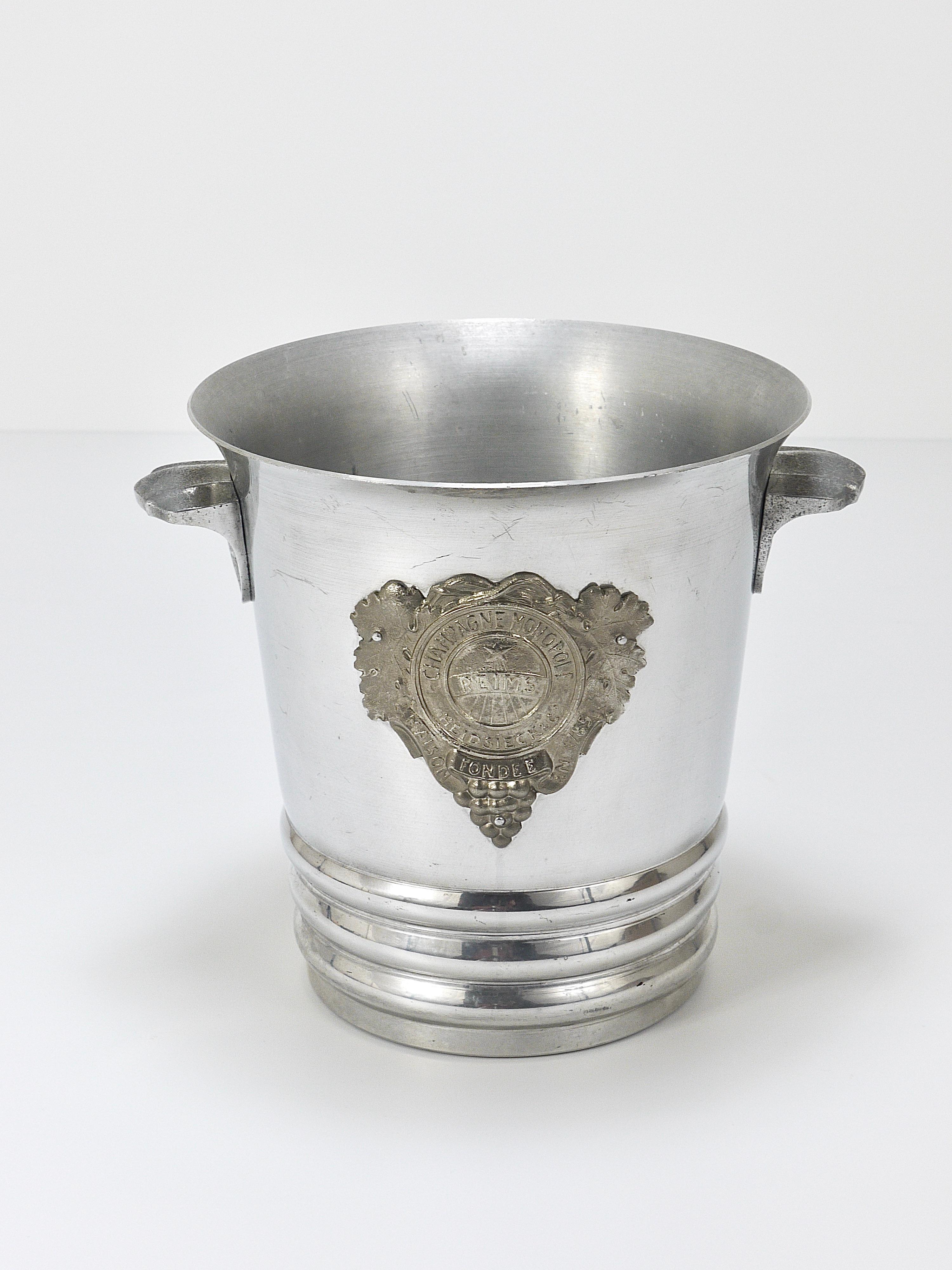 A beautiful Art Deco Champagne bucket from the 1930s / 1940s  by Champagne Monopole Heidsieck & Co. Made of polished aluminum with two nice handles and a decorative badge with  grapevines  and Heidsieck & Co lettering on its front. The handles and