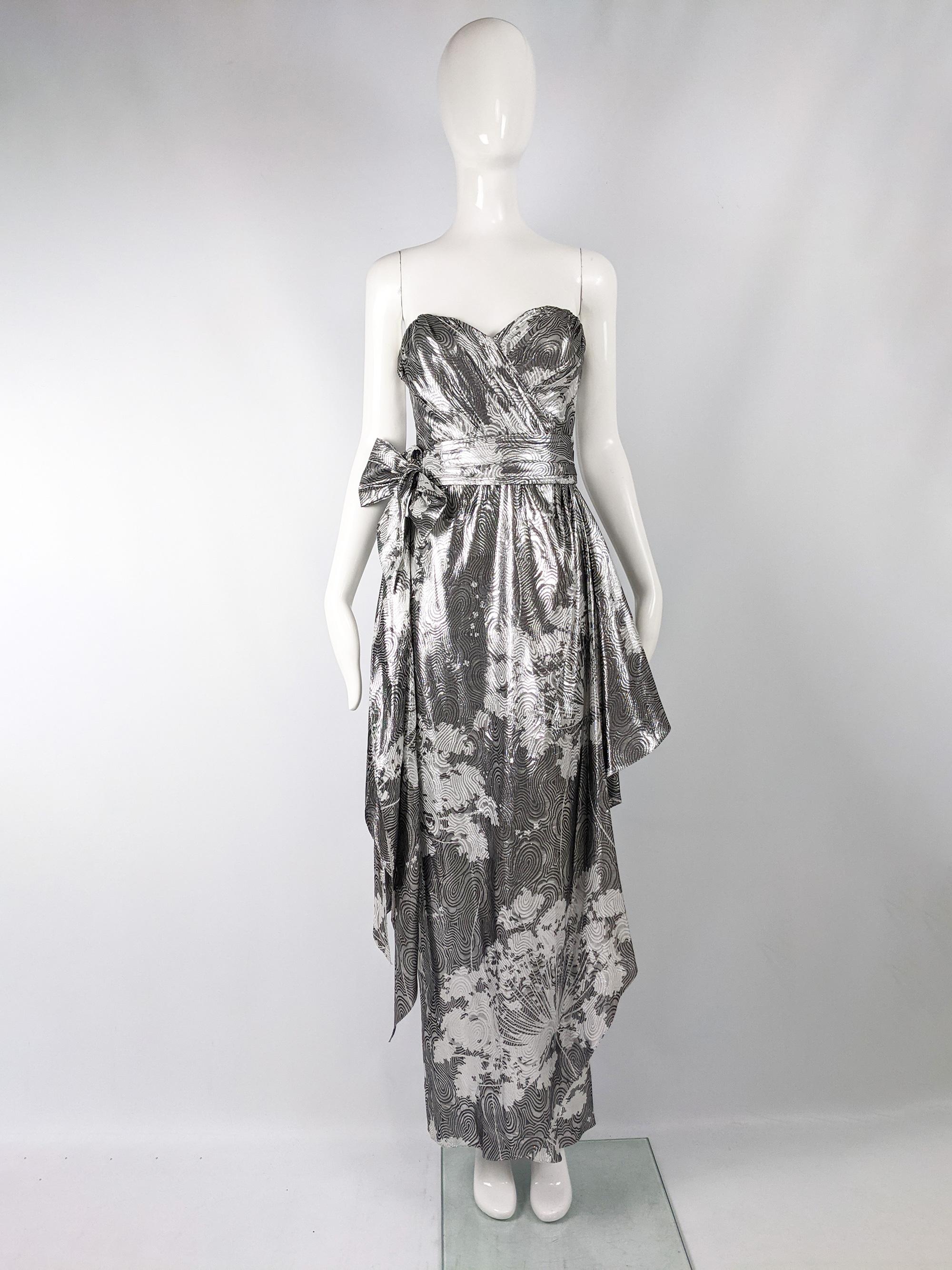 An elegant vintage evening gown from 1986 by British luxury designer Helen Anderson, it is made from a metallic silk lamé with a white floral print and shallow pattern throughout. It has a strapless design and a sash at the waist and drapes at the