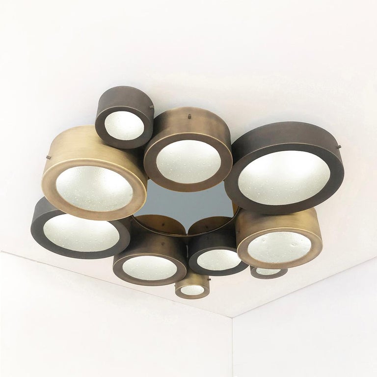 The Helios ceiling light features an imposing composition of illuminated Murano glass shades gravitating around a central tinted mirror. Shown as a flush mount in a three tone bronze finish, with a central gray blue mirror and our signature Murano