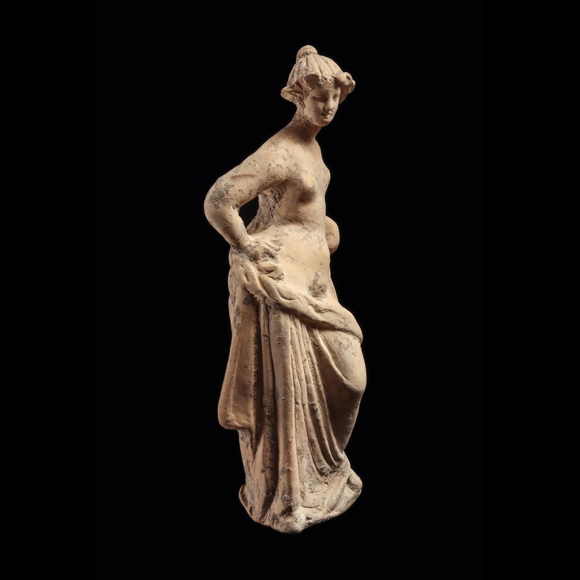 Terracotta statuette of a goddess, likely Aphrodite, leaning on a pillar. Her hair is pulled back into a bun, with framing locks around the forehead and an idealised face. Her left arm is resting on a pillar, right arm is on the hip, which is angled