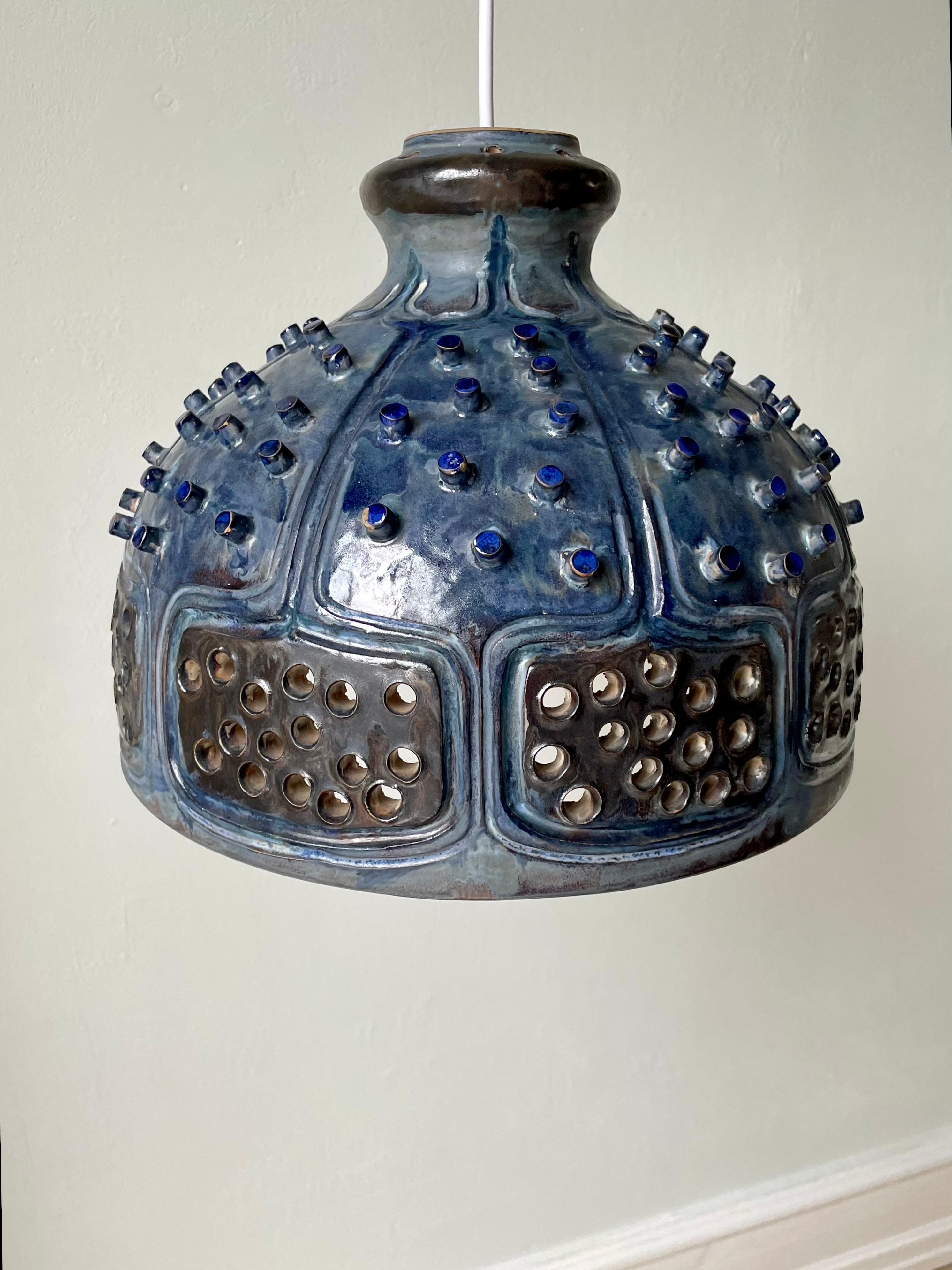 Blue monumental Brutalist stoneware pendant with intricate graphic and organic decorations. Handmade in the 1960s by ceramic artist Jette Hellerøe for Axella. Shiny petrol blue glaze over studded ornaments and soft graphic relief lines. Multiple