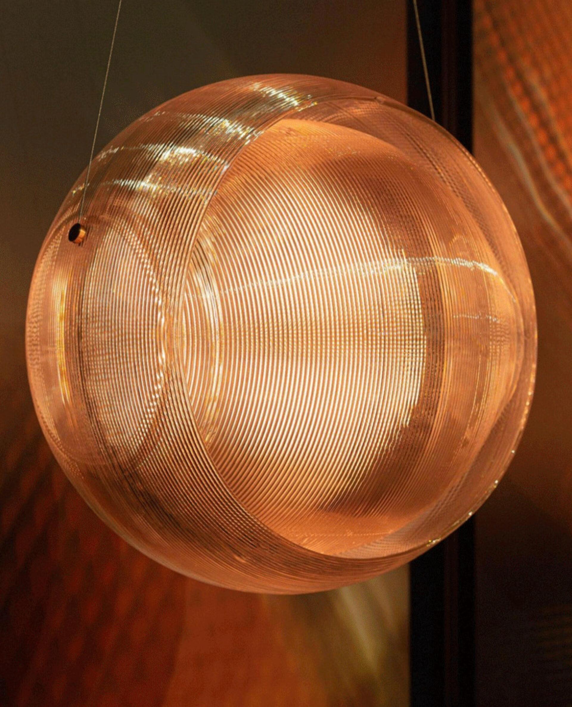 The Helm lamp designed by Kooij is a belted sphere grown additively from syrupy ribbons of molten, recycled plastic.
When passing through the corrugated structure, light breaks, softens and multiplies into an engulfing spread.