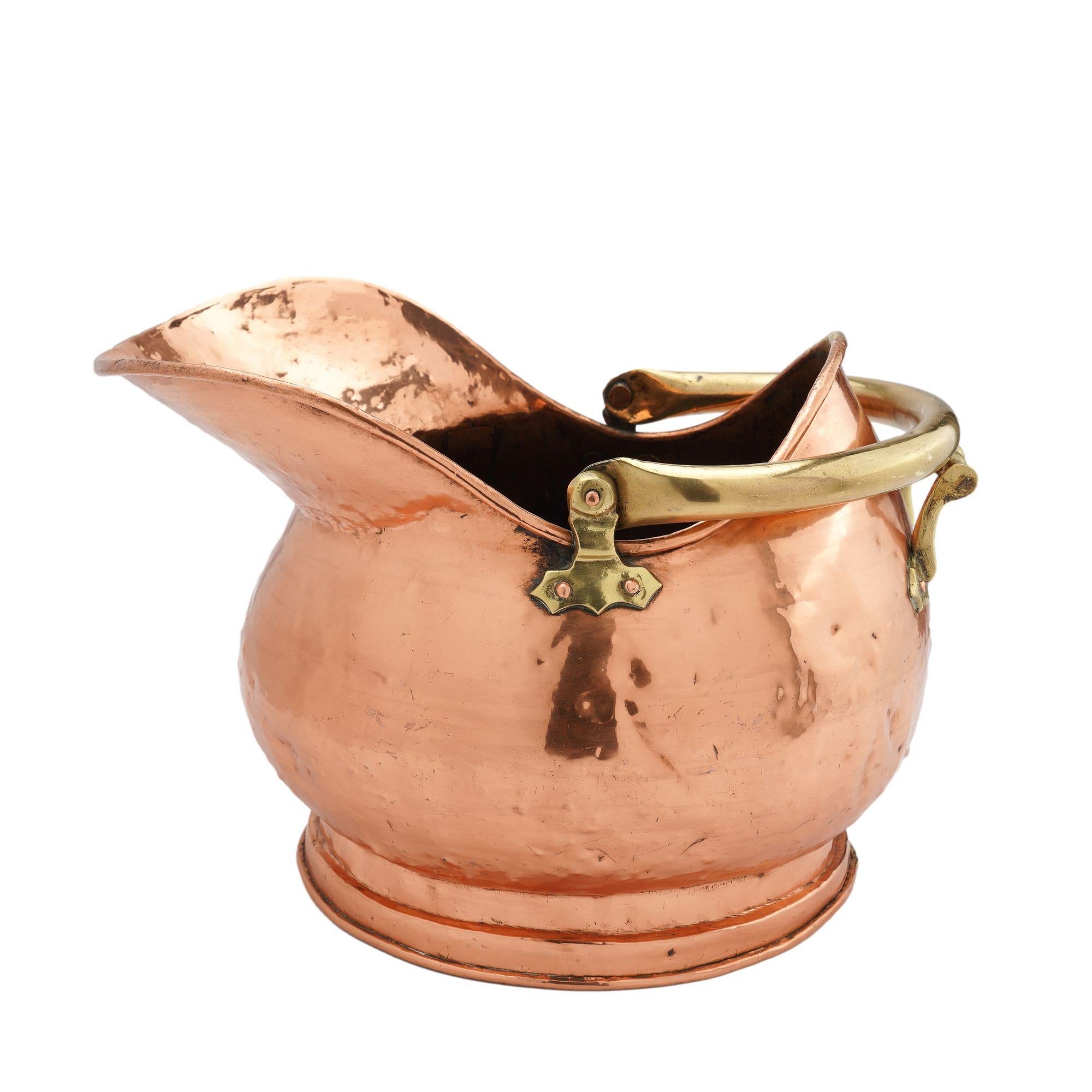 A generous bombé helmet shaped copper coal hod on a circular footed base. The hod is fitted with a brass bail handle mounted on cast brass pivot fittings and a cast brass tilter grip on the back rim. The copper surface is hand hammered with dove